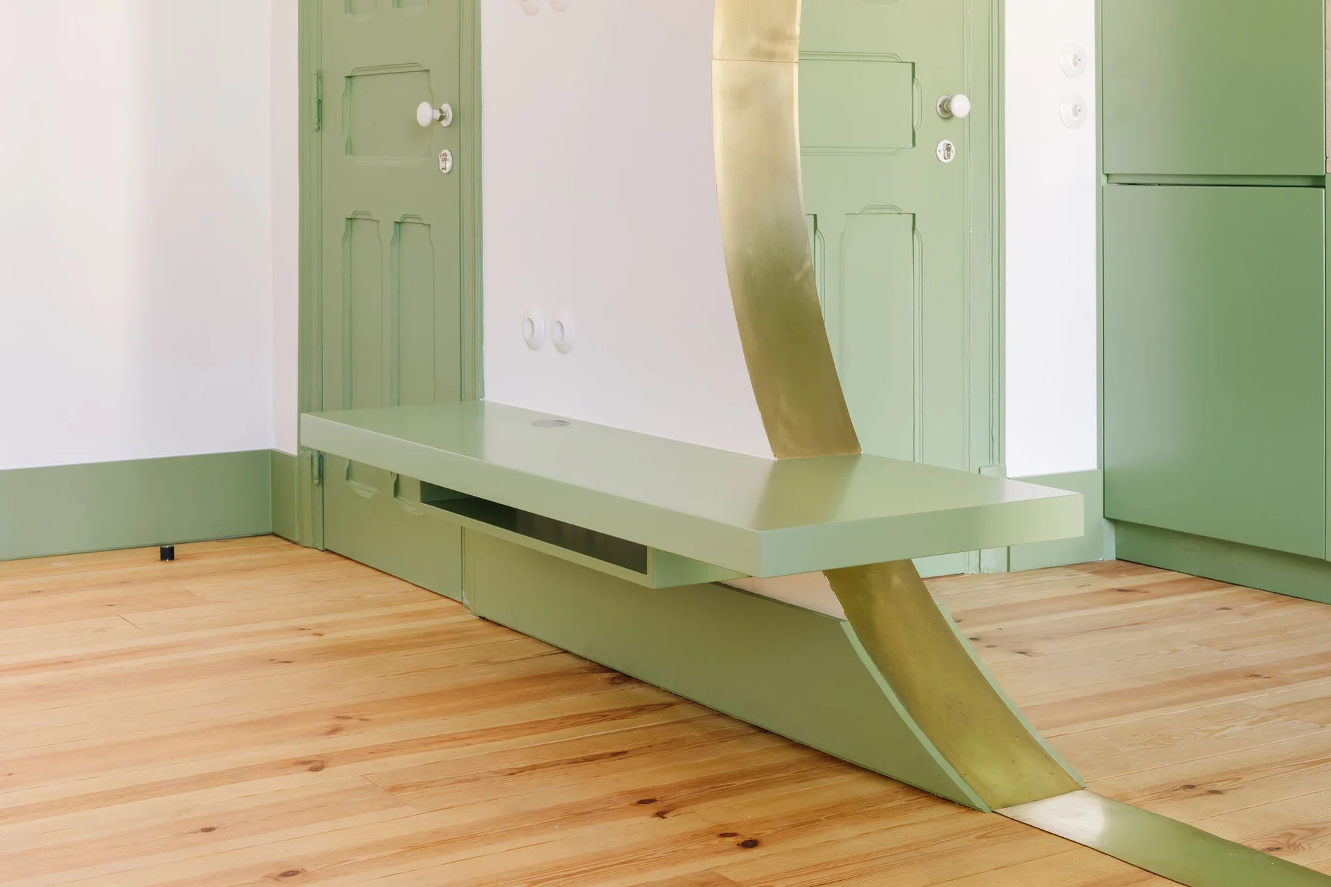 A minimalist hallway features a sage green painted built-in bench with a curved brass accent flowing down to the light wood plank floor.