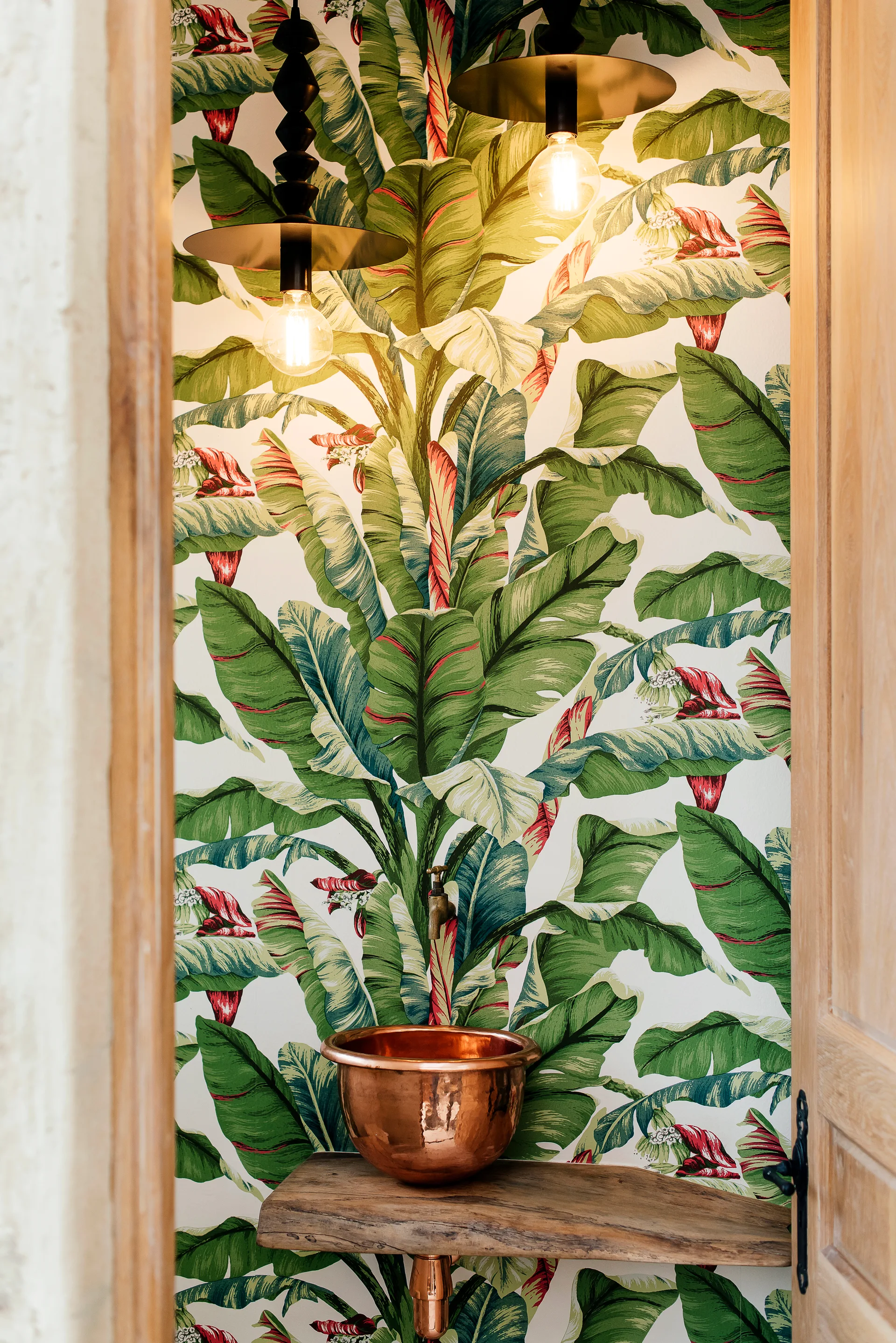 A small powder room features green banana leaf wallpaper, a copper vessel sink on a live-edge wood shelf, and two pendant lights.