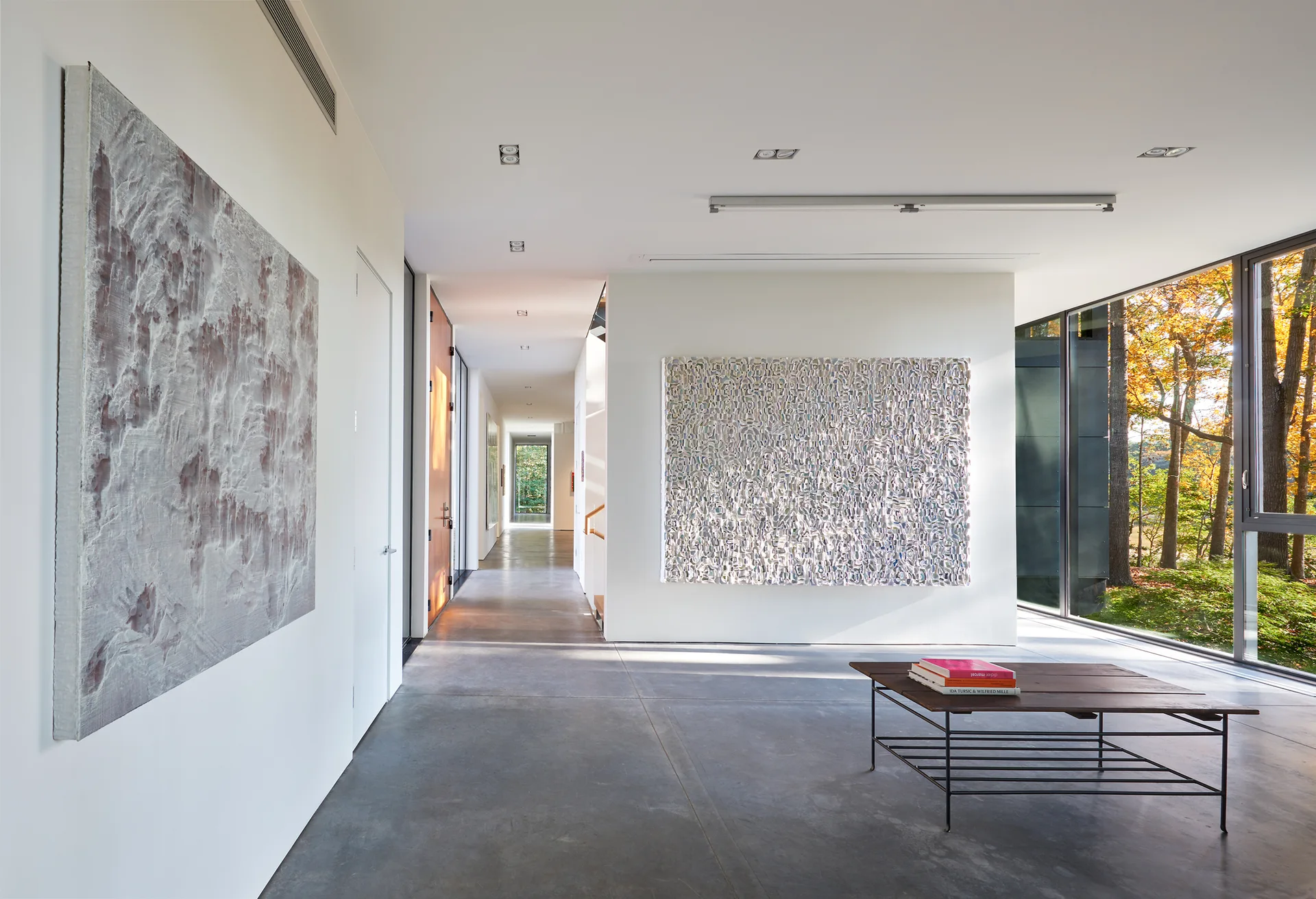 A wide minimalist hallway featuring smooth grey concrete flooring, white walls with large abstract paintings, and a wall of windows showing trees.