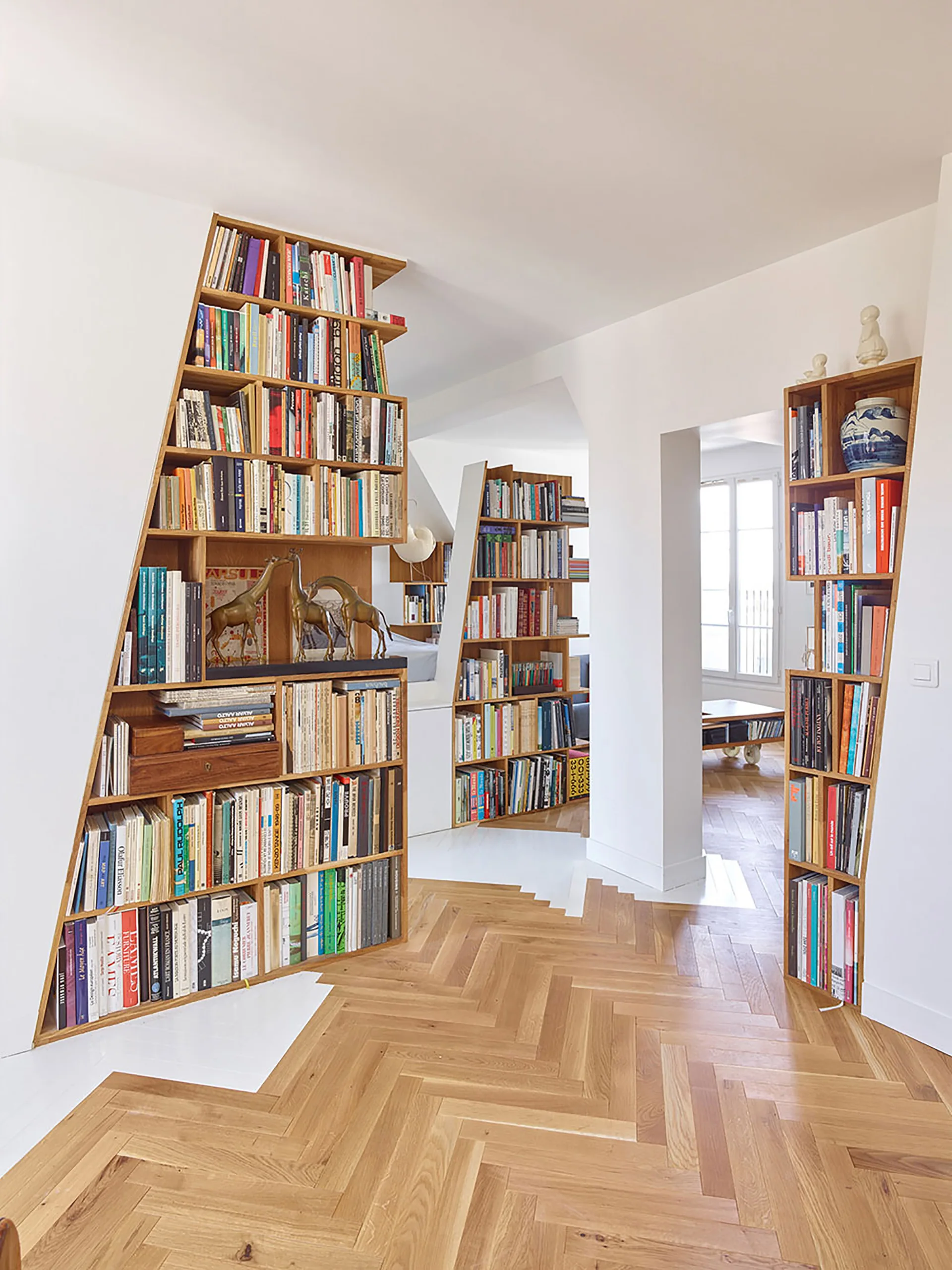 Custom wooden bookshelves with slanted profiles sit on a herringbone wood floor in a bright room with white walls.