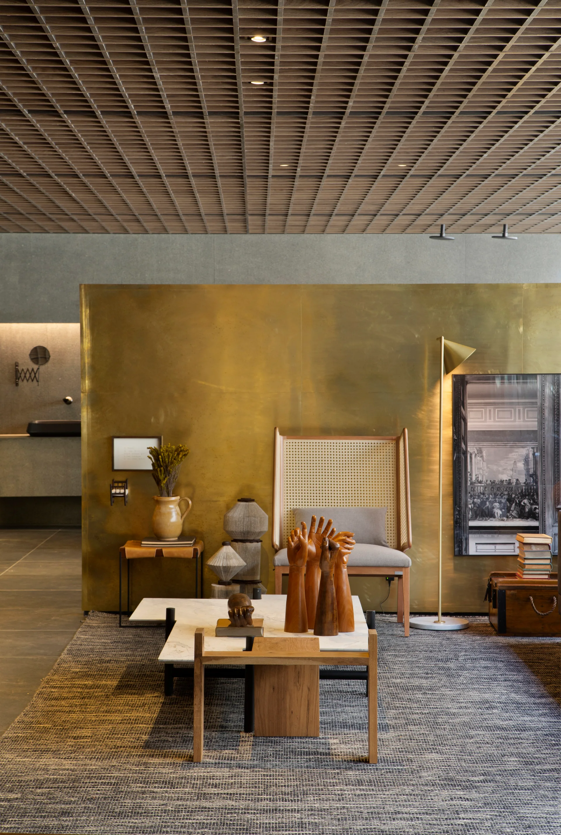A modern lounge featuring a high-back cane chair, a low marble coffee table, and a large brass partition wall beneath a wooden grid ceiling.