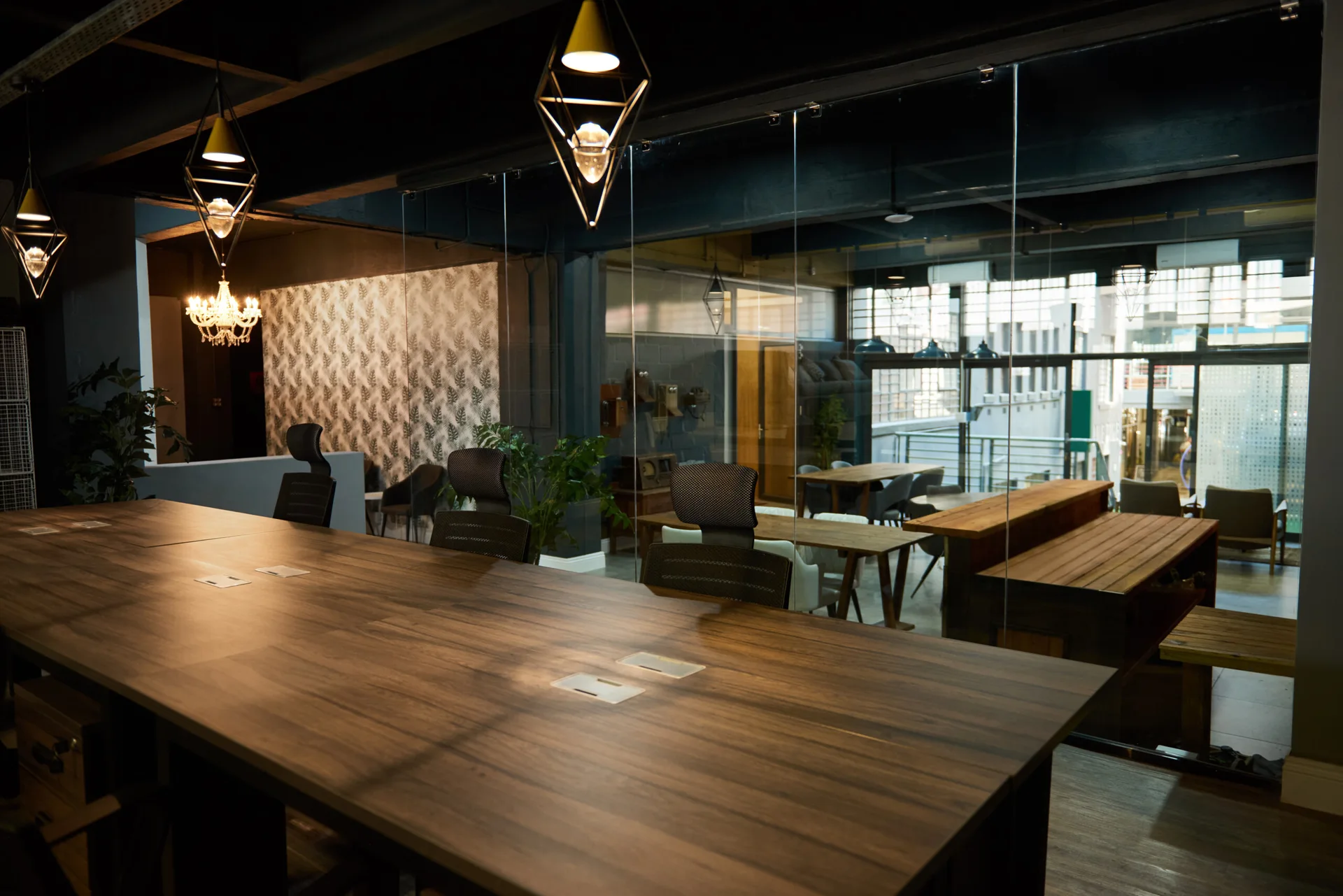 An industrial workspace featuring a long wooden conference table, black ergonomic chairs, and full-height glass partitions under a dark ceiling.