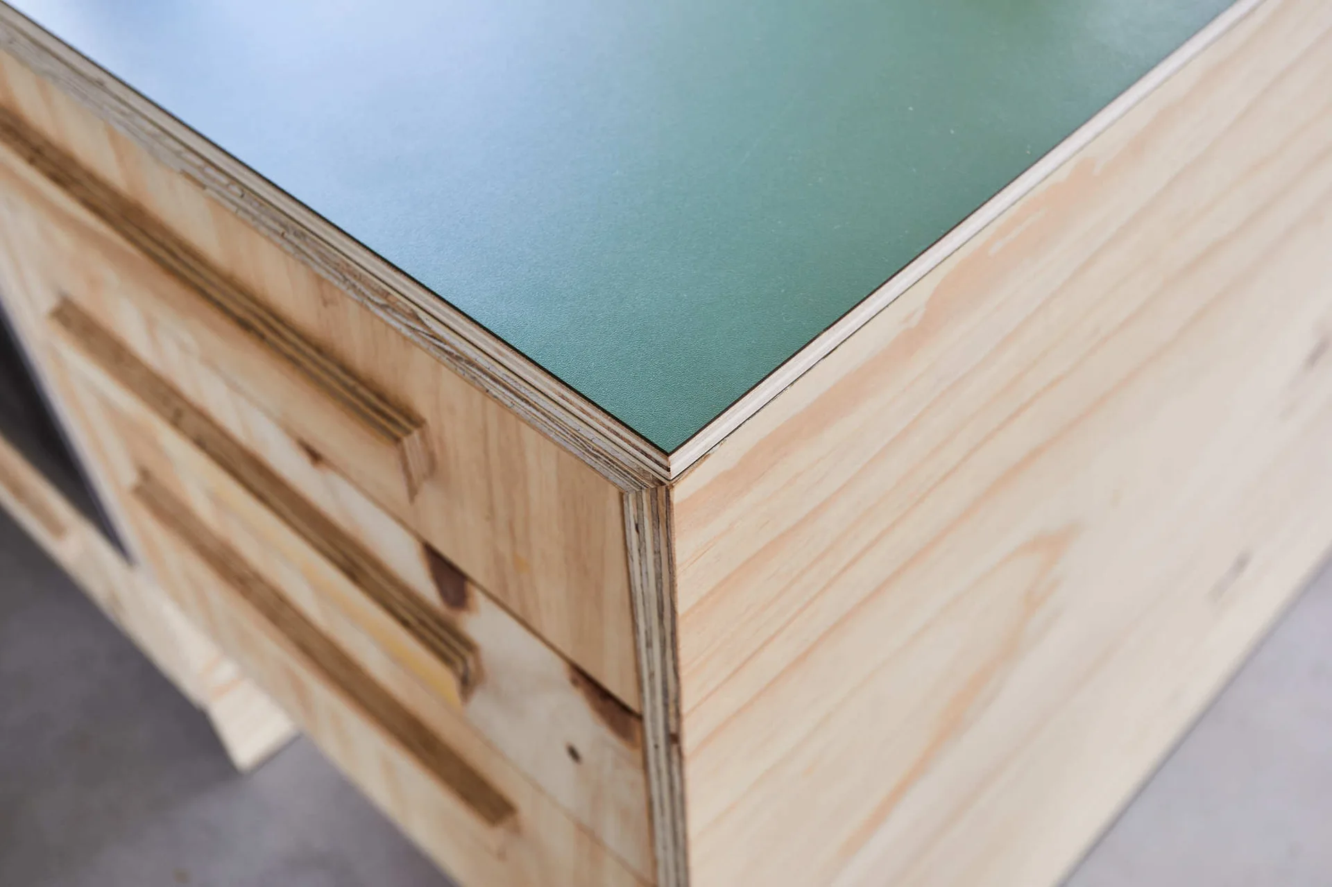 A close-up of a custom plywood cabinet featuring exposed layered edges, drawers with integrated wood pulls, and a matte green laminate top.