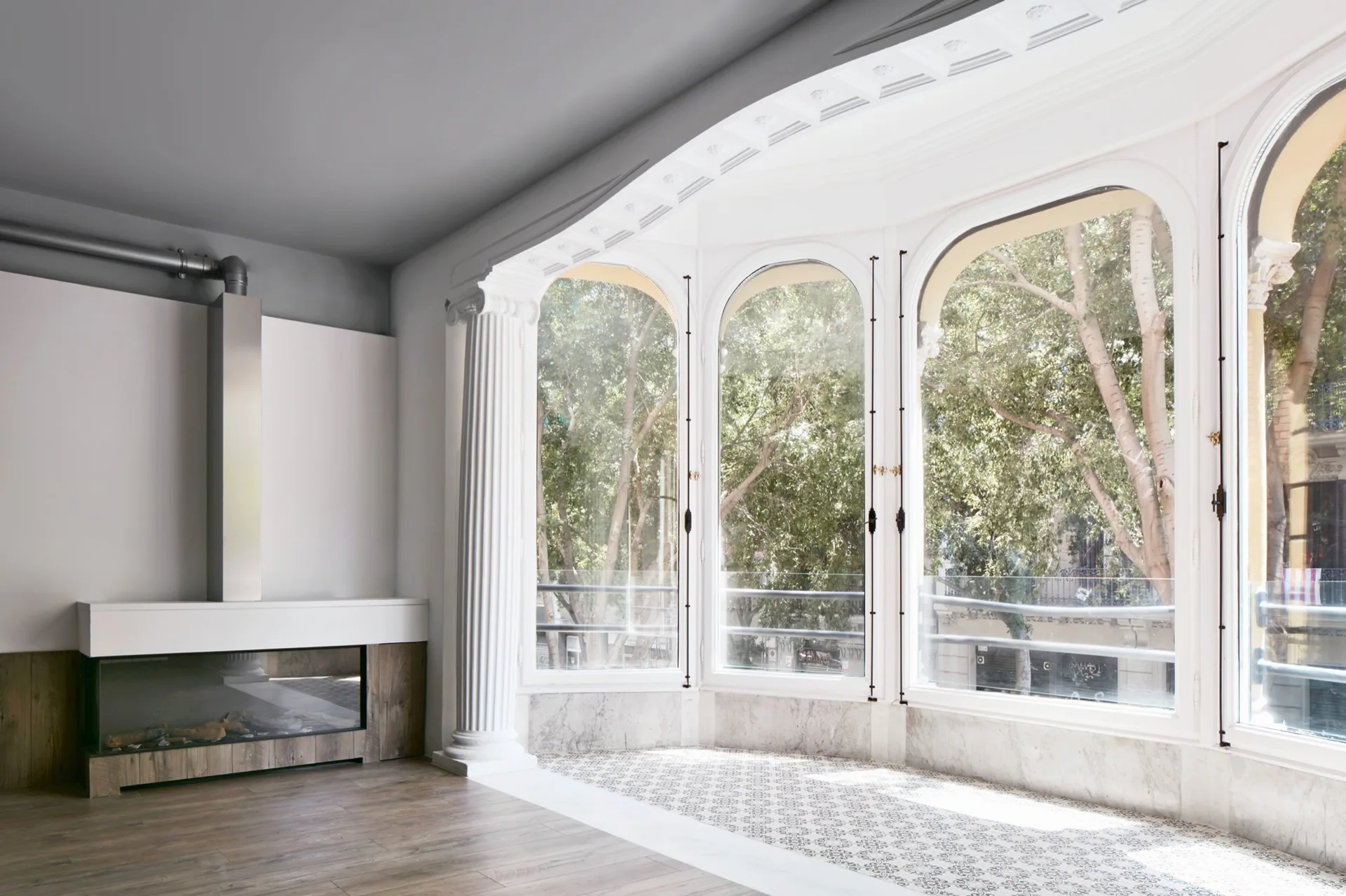 An empty room features large arched bay windows framed by a classical fluted column, patterned floor tiles, and a modern fireplace.