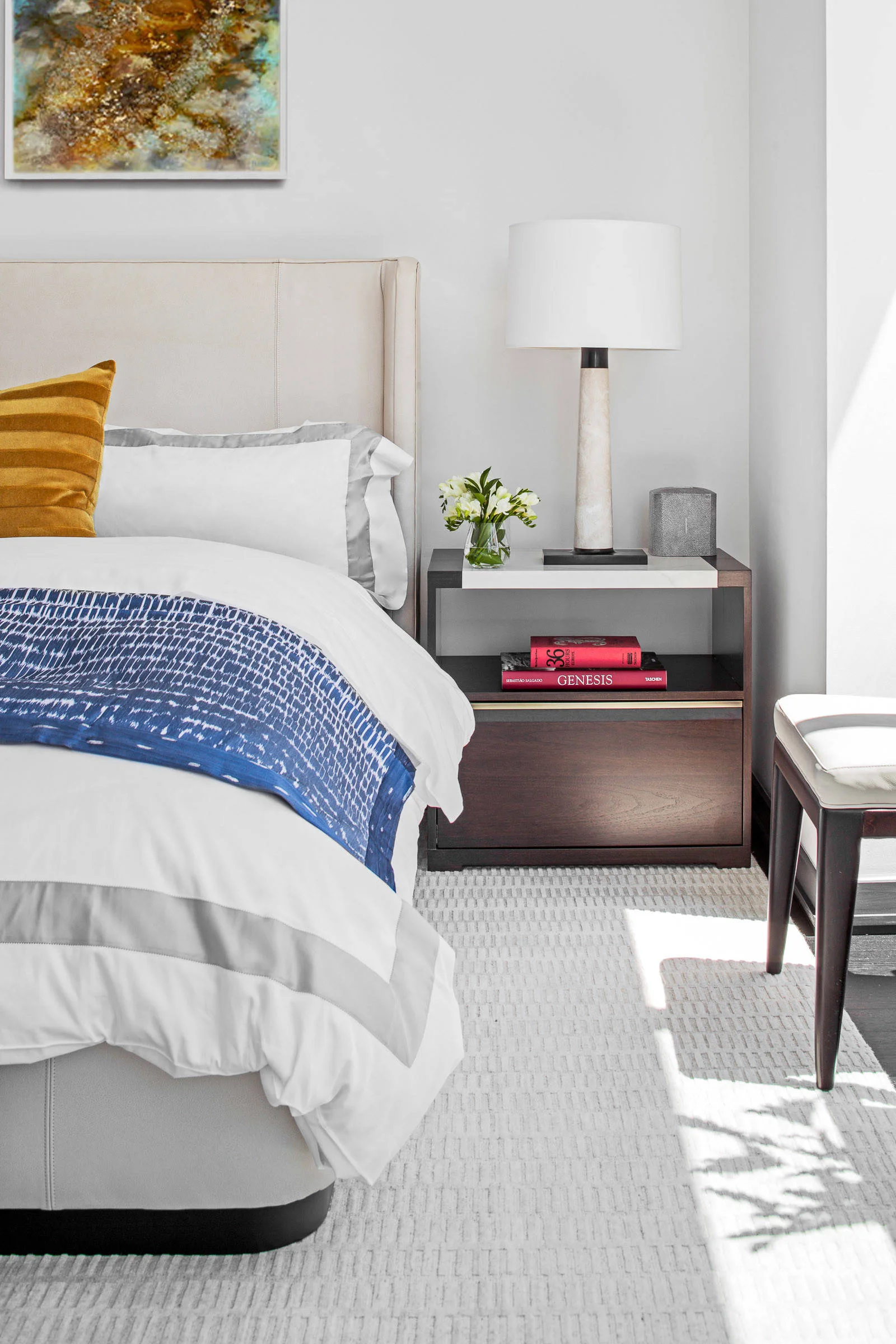 A modern bedroom features an upholstered bed with a blue patterned throw, a dark wood nightstand with a marble top, and a stone table lamp.