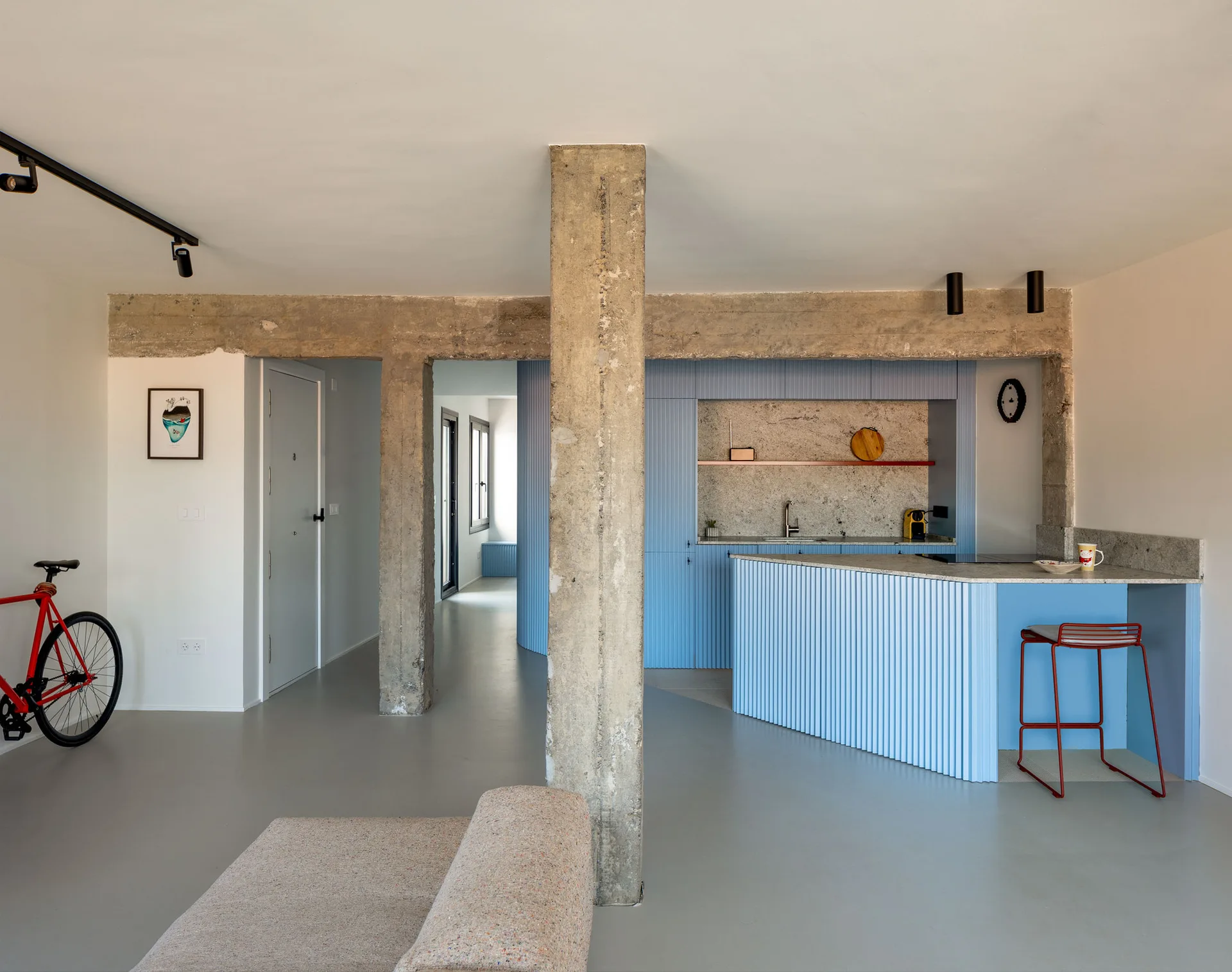 An open-concept kitchen features blue ribbed cabinets, an island with a stone countertop, a red stool, and exposed concrete structural pillars.