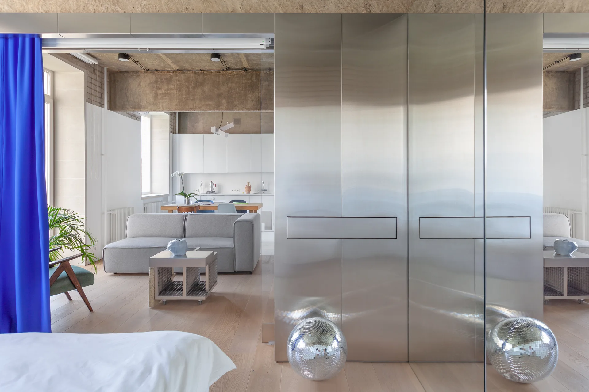 A modern apartment featuring a large brushed stainless steel partition wall, raw concrete ceiling, light wood floors, and a bright blue curtain.