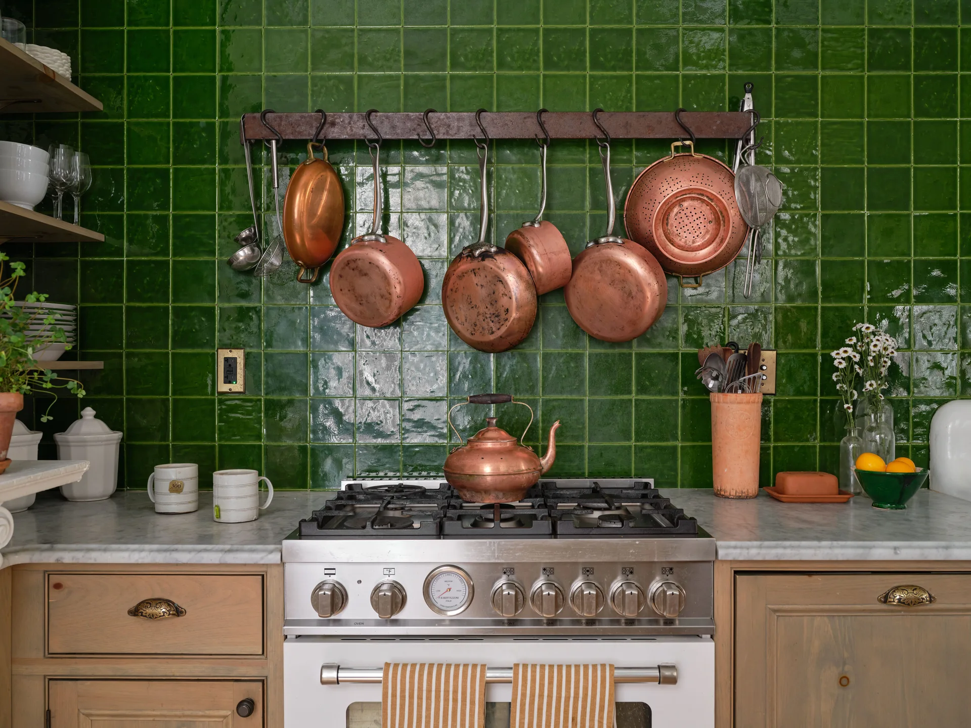 A stainless steel kitchen range stands before a glossy green tiled backsplash where copper pots and pans hang from a rustic metal rack.