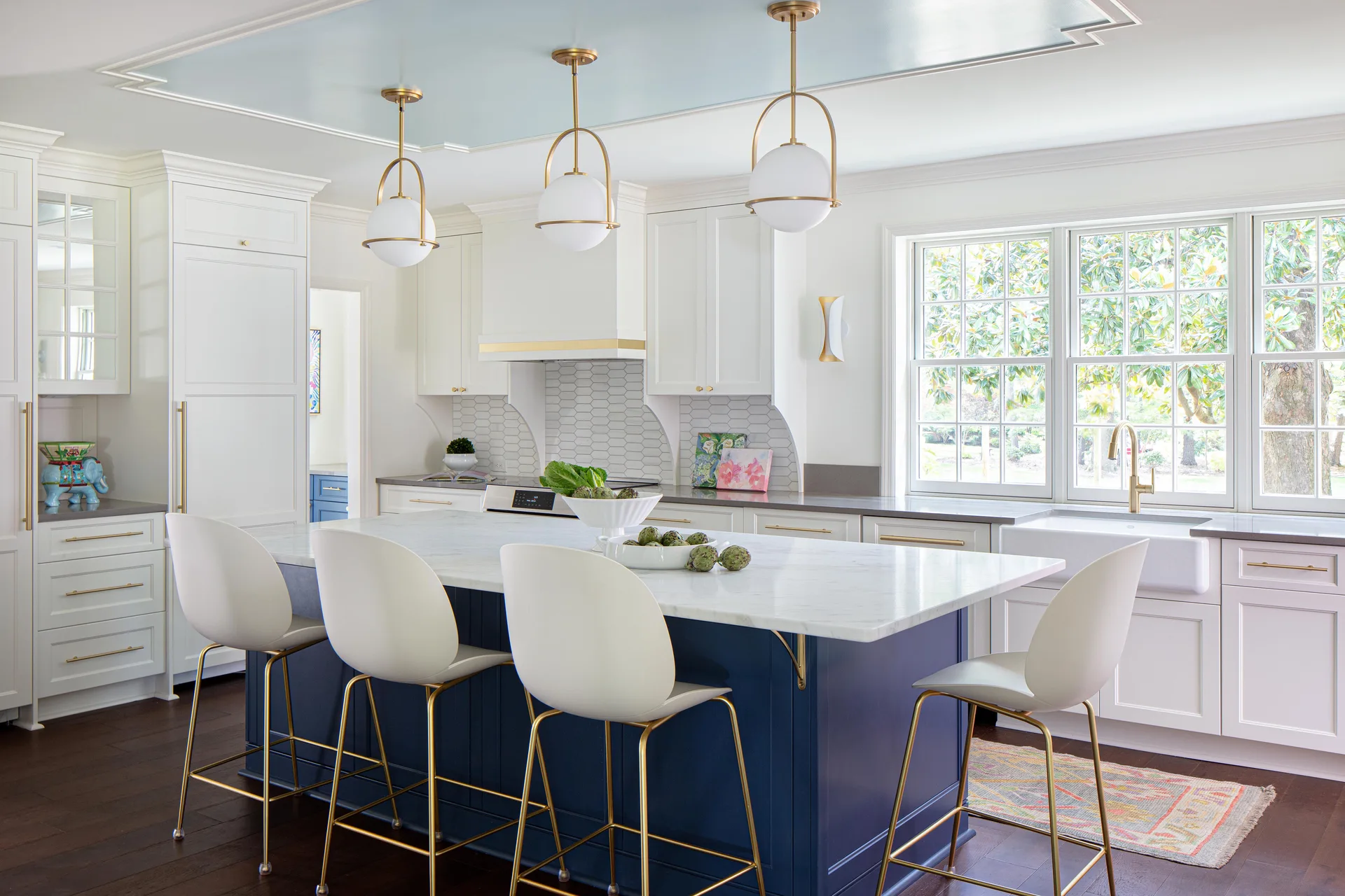 A bright transitional kitchen with a navy blue island, white marble countertops, three gold globe pendant lights, and a light blue painted tray ceiling.