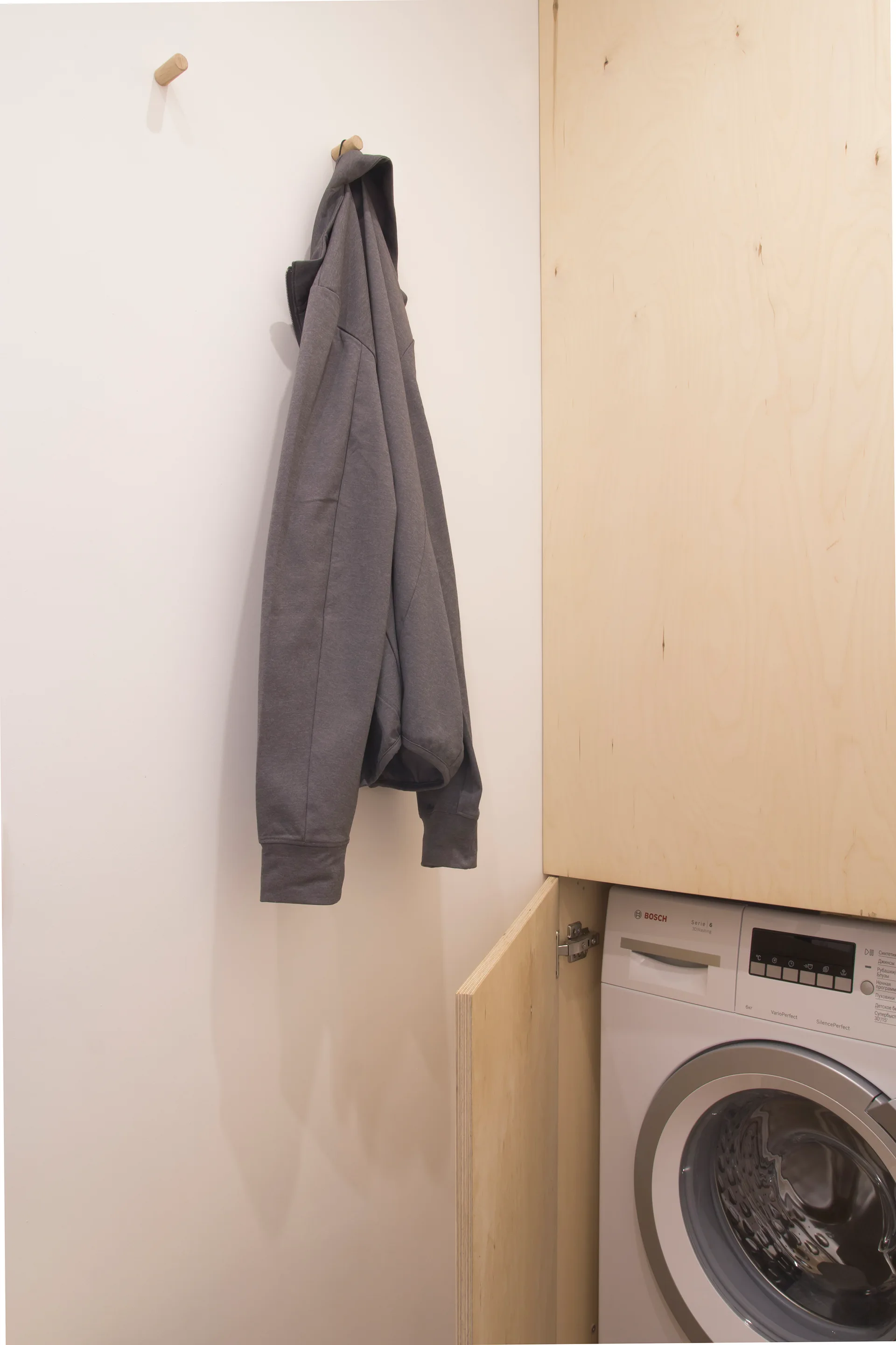 A white washing machine sits partially concealed inside blonde wood cabinetry next to a plain white wall with wooden coat pegs.