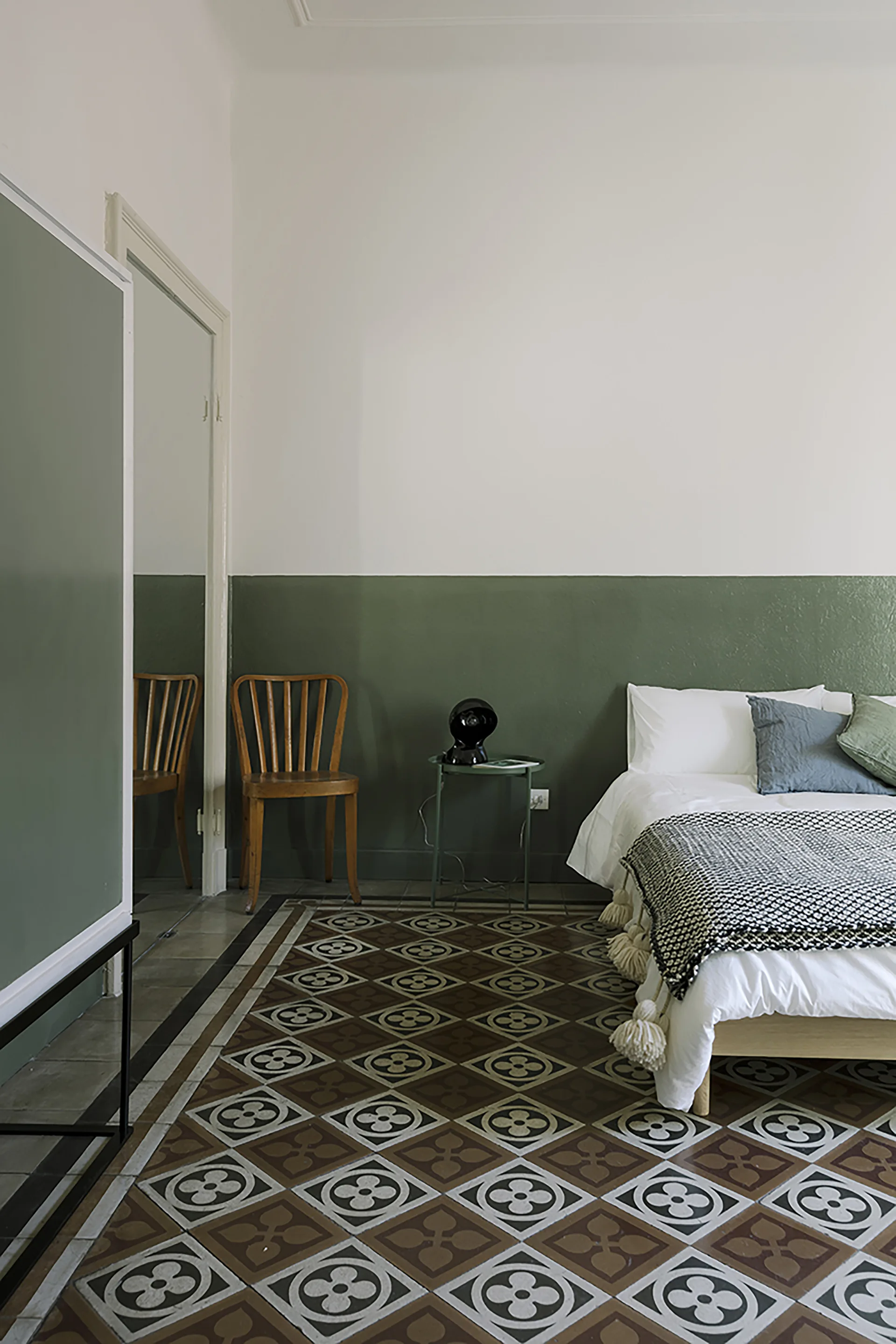 A bedroom features a bed with white linens, a wooden chair, a green color-blocked wall, and geometric brown and white floor tiles.