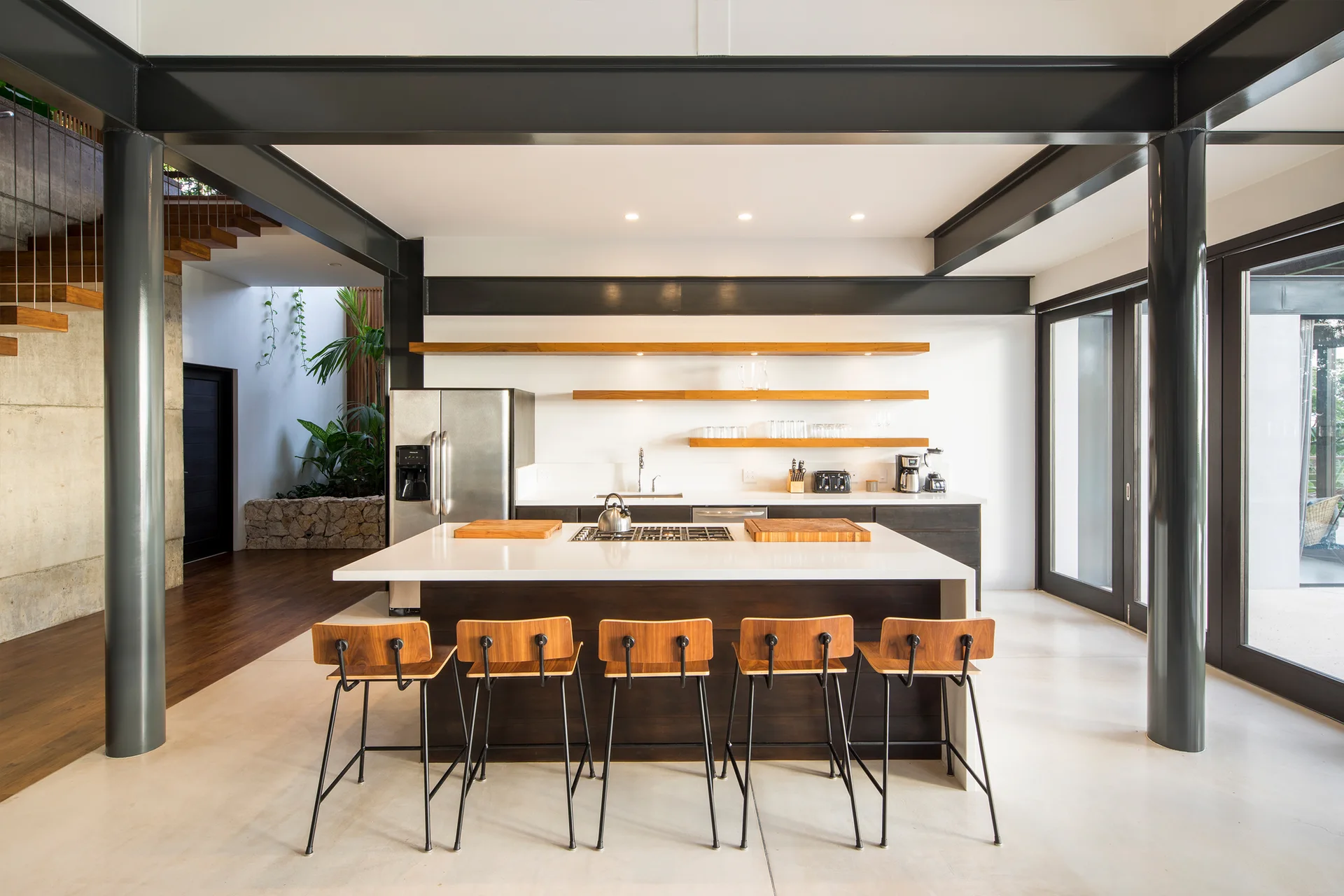 A modern industrial kitchen features a large white island with wooden bar stools under exposed black metal beams and floating wood shelves.