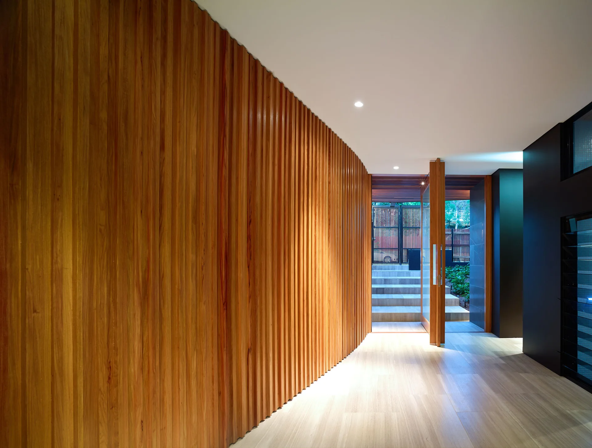 A modern entryway hallway featuring a large vertical wood slatted accent wall, light stone tile floors, and an open pivot glass door leading outside.
