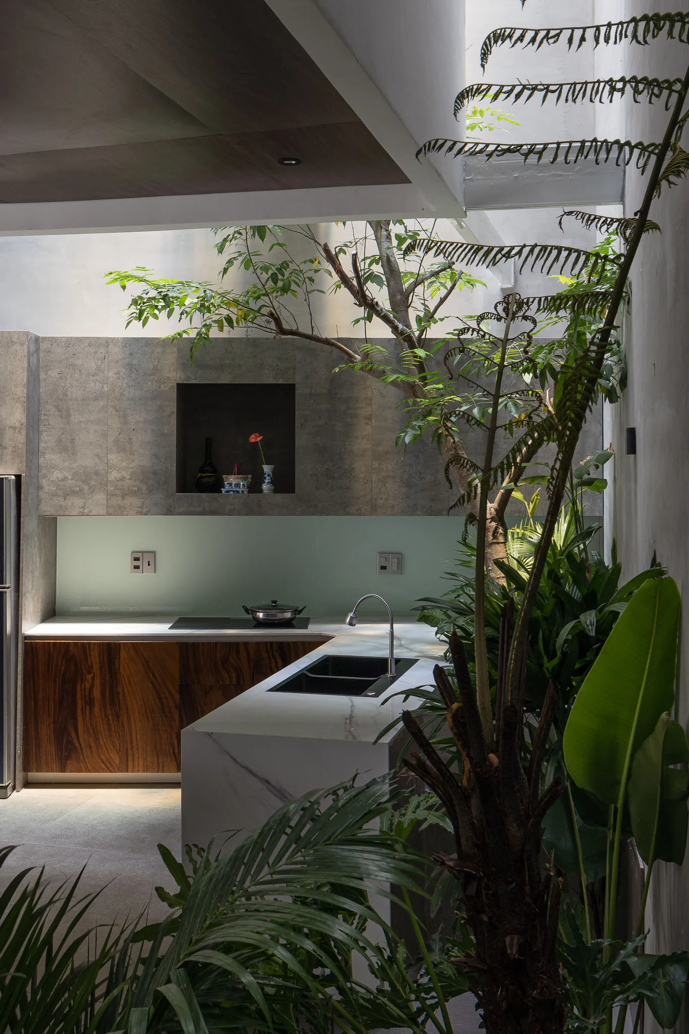 A modern kitchen features wood-grained lower cabinets, a marble-patterned island, and an indoor tree growing beneath a bright overhead skylight.