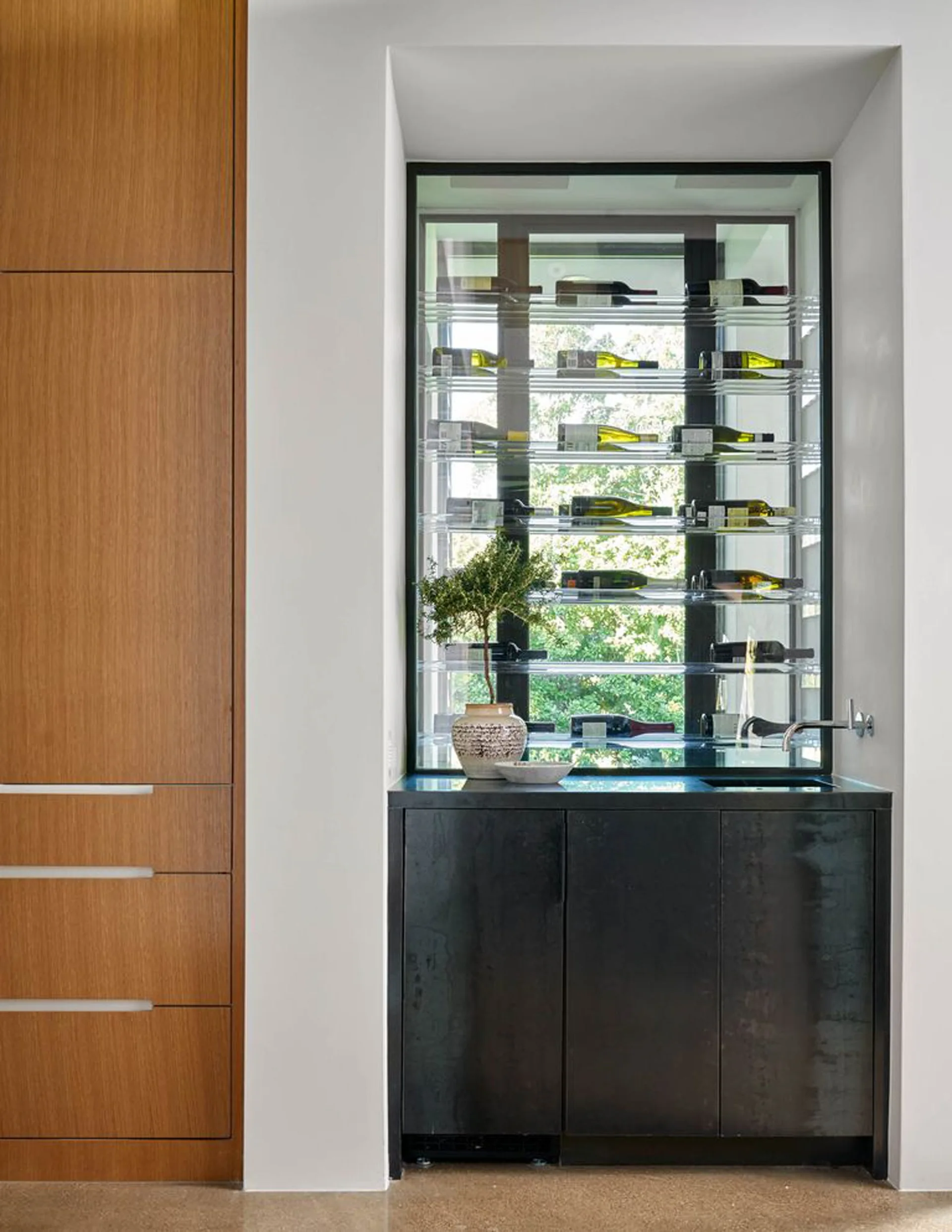 A recessed wet bar features a dark metal lower cabinet, a small sink, and a transparent glass wine rack set against a window with outdoor views.