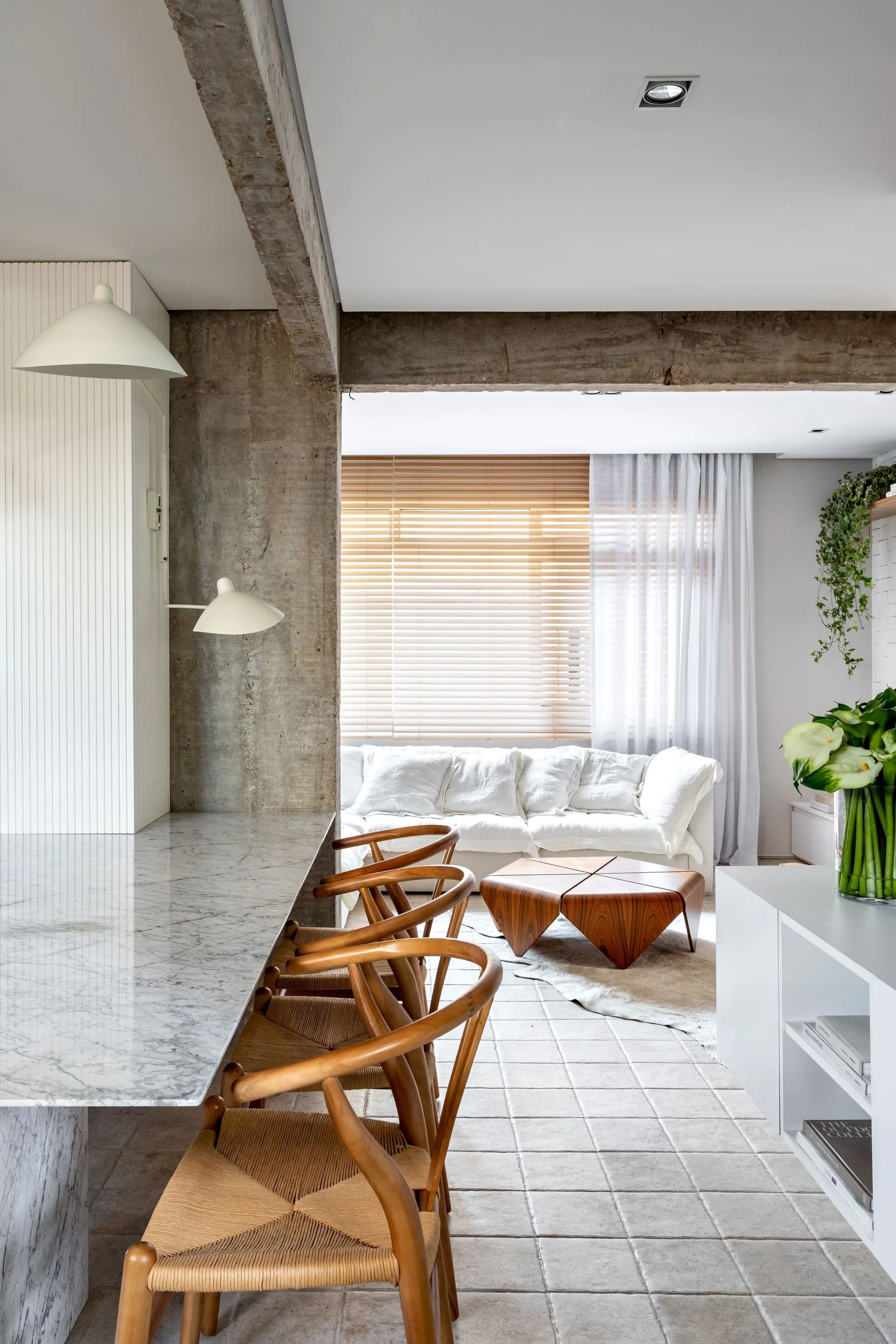 An open-plan interior featuring a white marble dining counter with wooden bar stools, an exposed concrete beam, and a white sofa in the background.