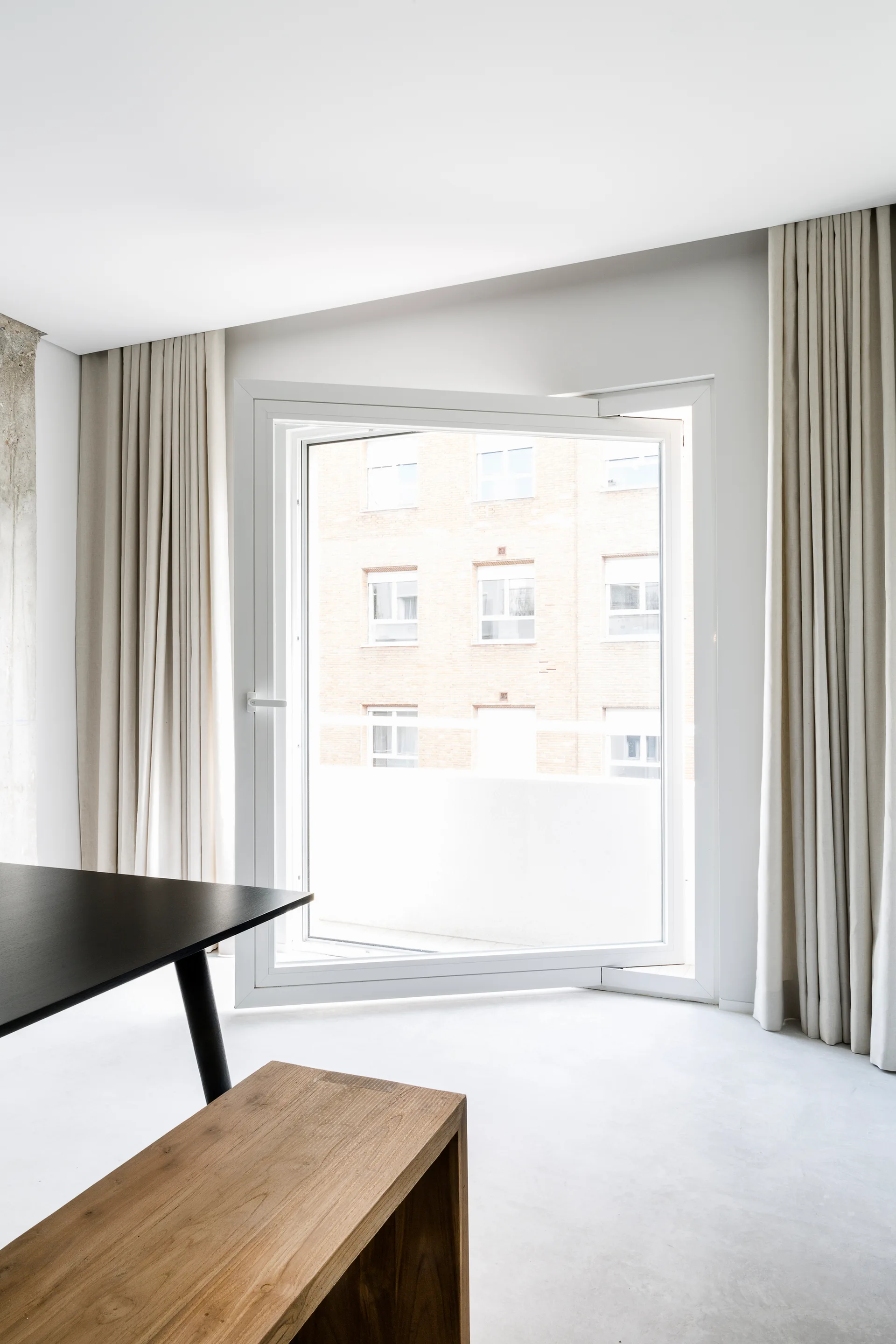 A minimalist dining area featuring a black table, a wooden bench, and a large white pivot window framed by beige floor-to-ceiling curtains.