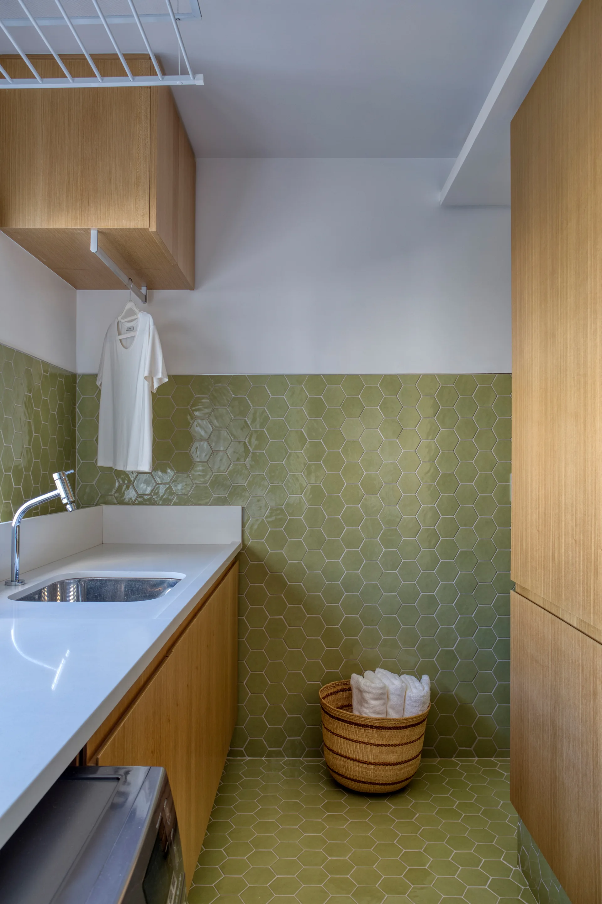 A modern laundry room featuring a white countertop with an undermount sink, light wood cabinetry, and sage green hexagonal floor and wall tiles.