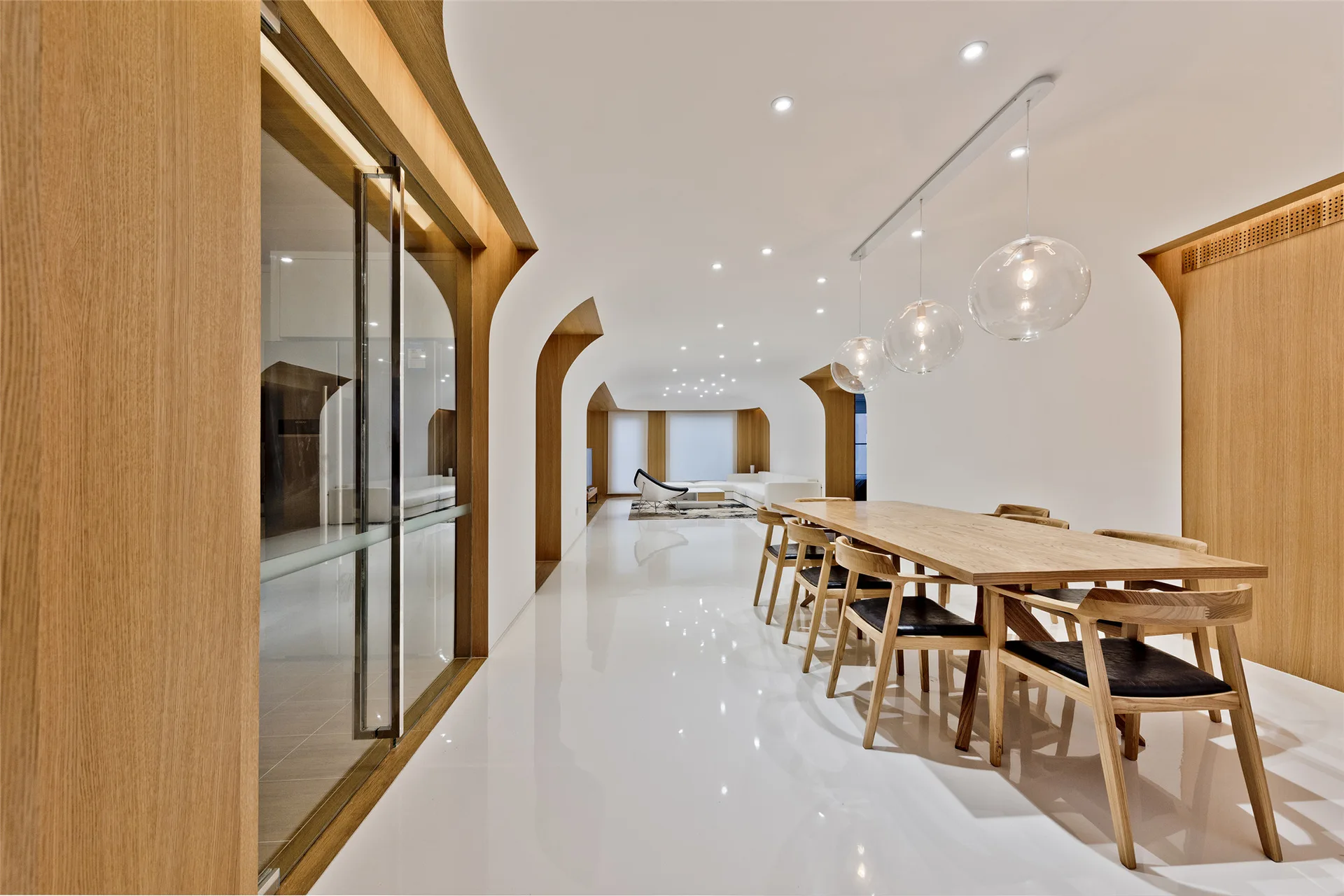 A long wooden dining table and chairs sit on reflective white floors beneath glass globe pendant lights and architectural wooden arches.