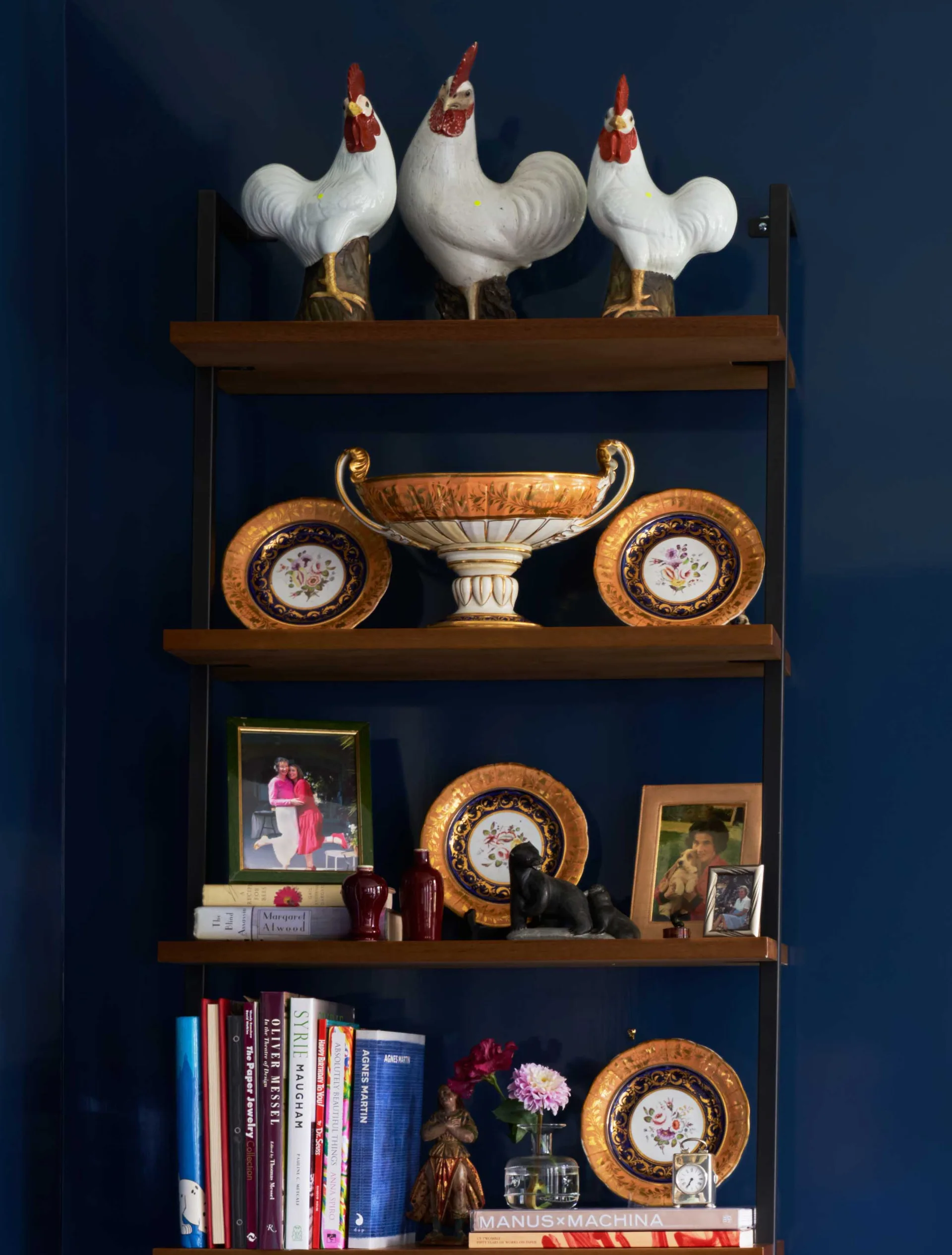 A dark wood and metal open shelving unit mounted on a deep navy blue wall, styled with rooster figurines, gold decorative plates, and books.