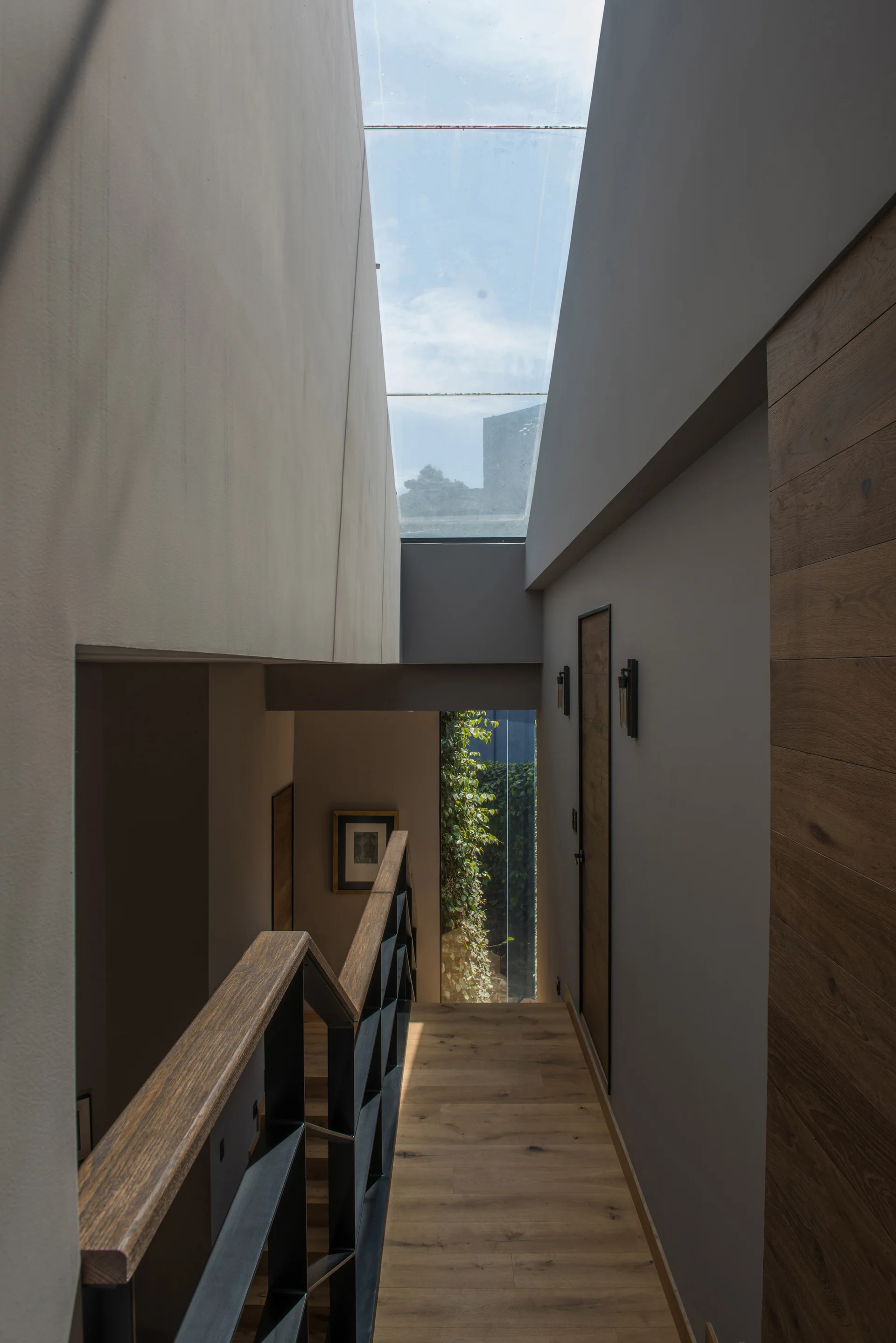 A modern staircase features light wood steps, a black geometric metal railing, grey walls, and a large skylight letting in natural light.
