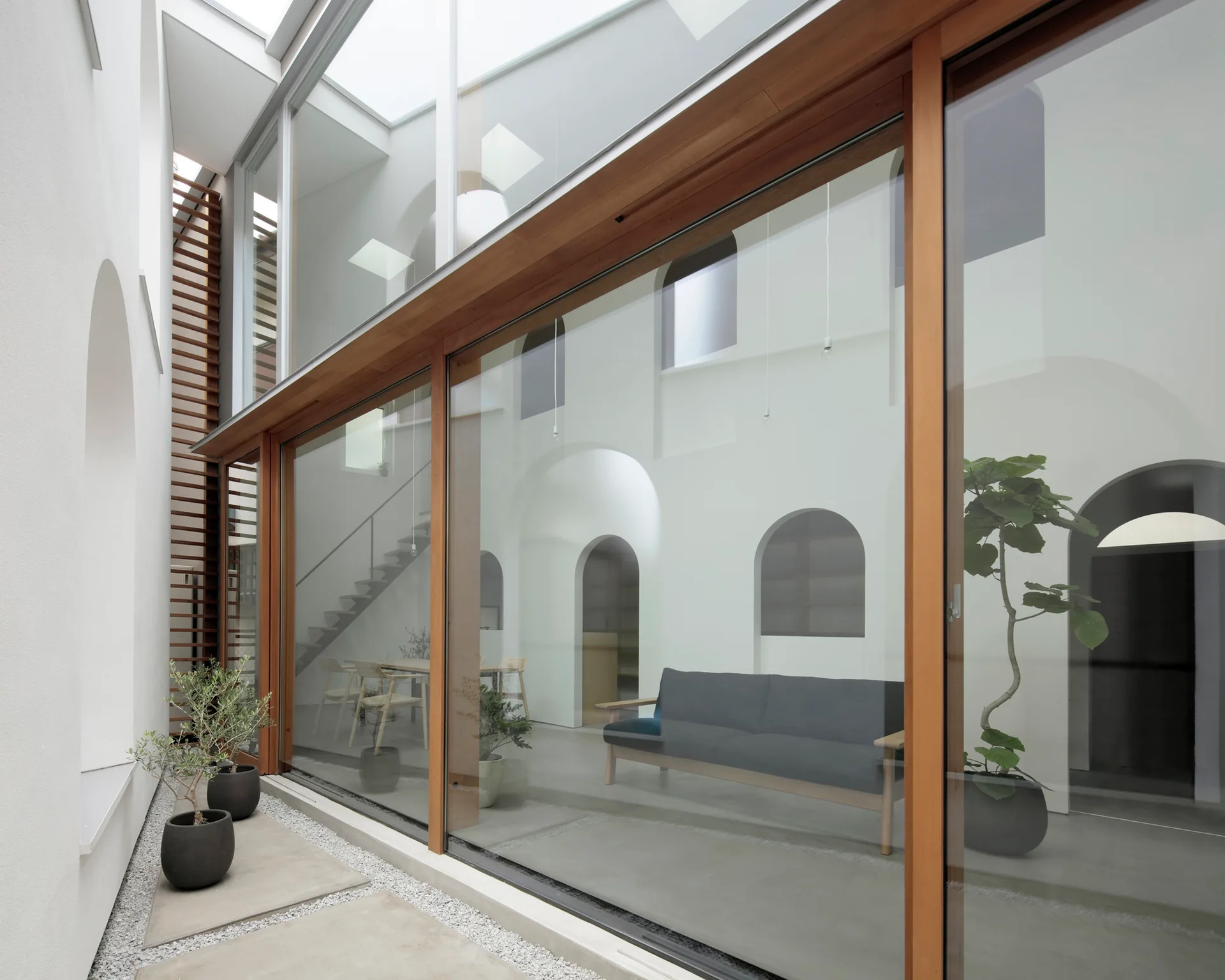 A minimalist interior with white walls and arched openings is visible through a large wood-framed glass facade overlooking a gravel courtyard.