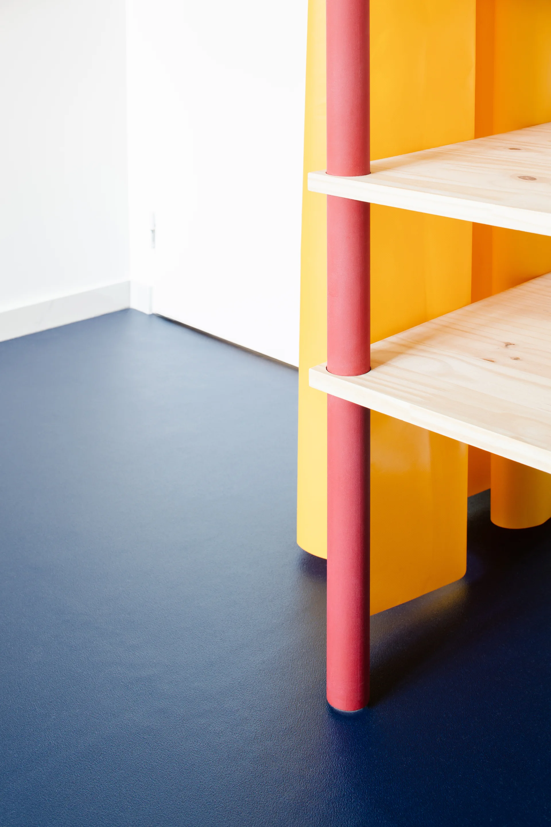 Close-up of a light wood shelving unit with a red cylindrical support post and yellow vertical divider on a dark blue floor.