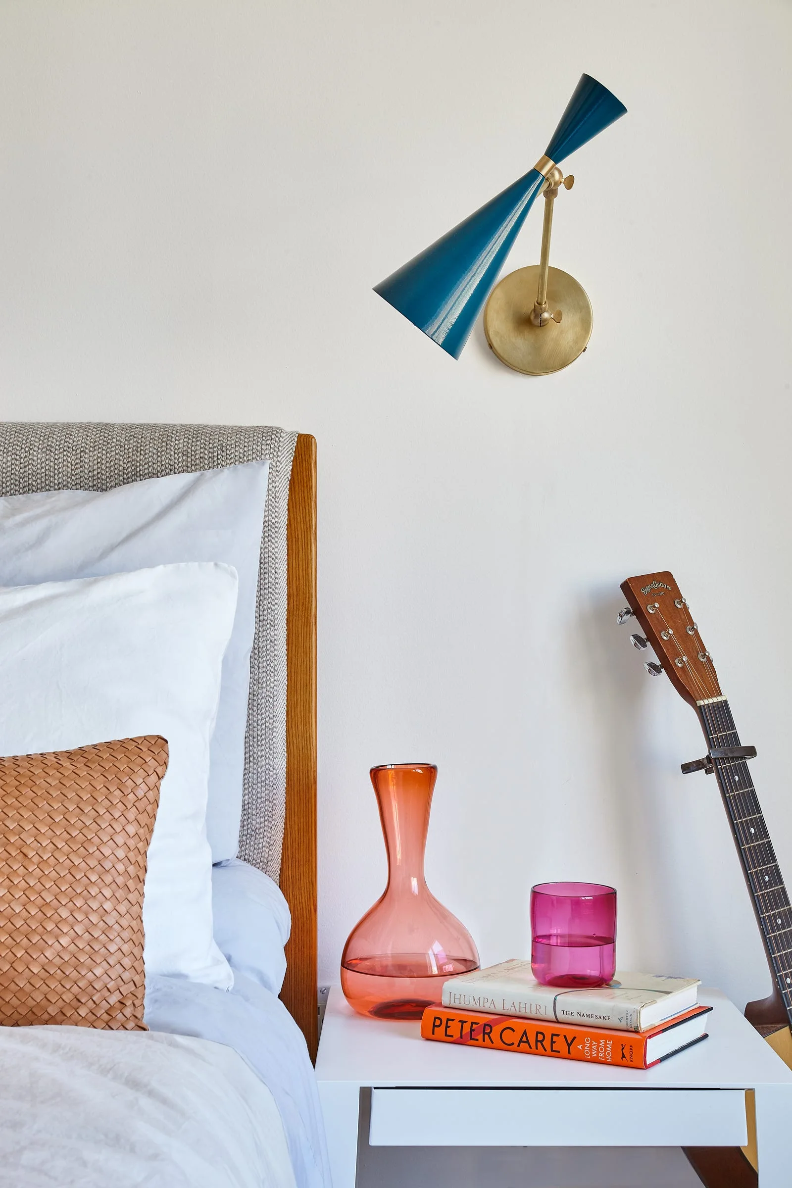 A white nightstand holding books and colorful glassware sits next to a bed under a blue cone-shaped wall sconce.