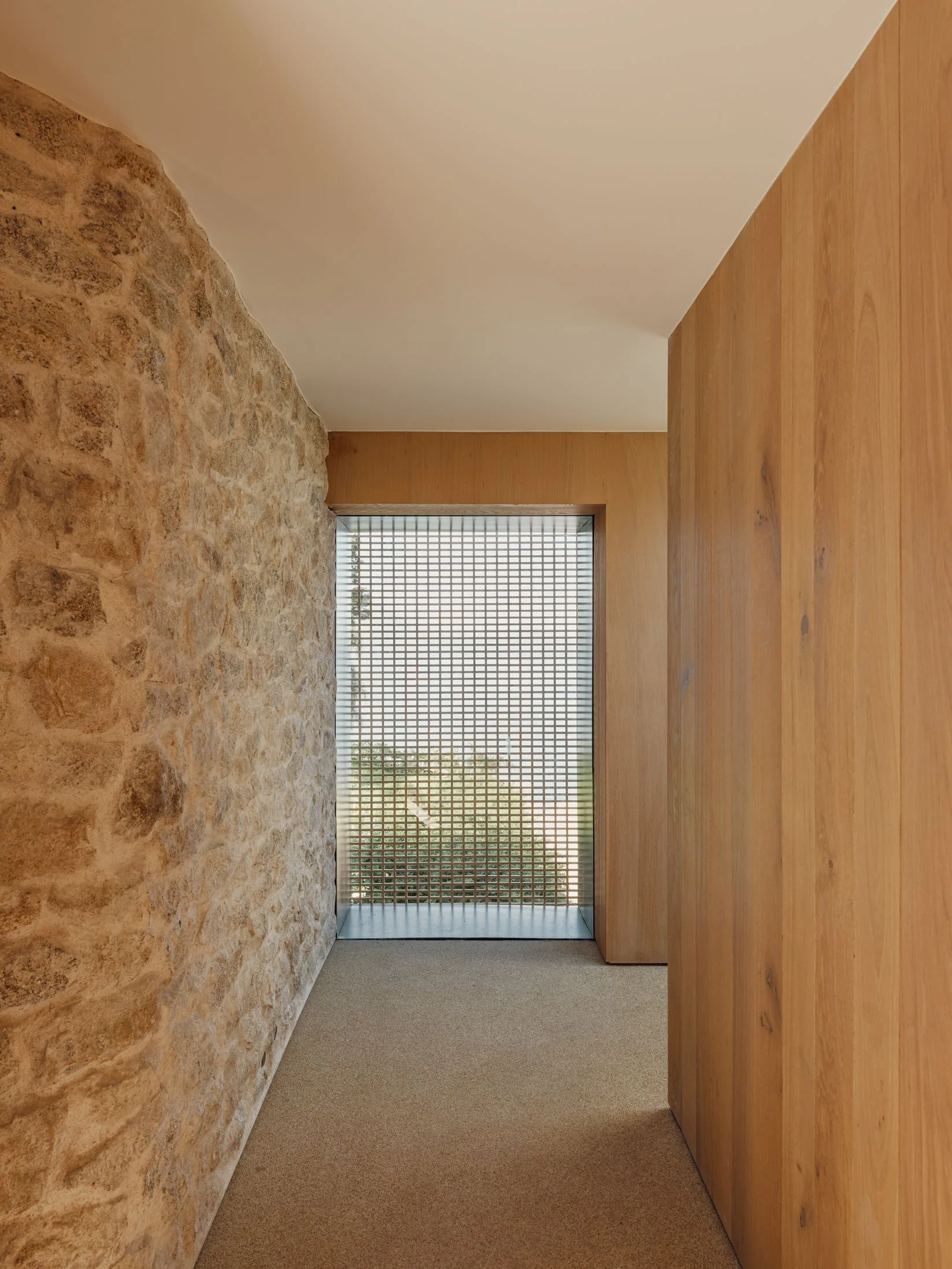 A minimalist hallway featuring a rough textured stone wall on the left, smooth timber panels on the right, and a gridded glass window at the far end.