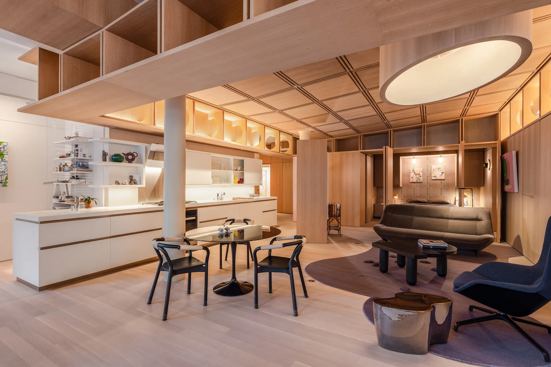 An open-concept living room and kitchen features a light wood coffered ceiling, wood paneling, white cabinets, and an organically shaped purple rug.