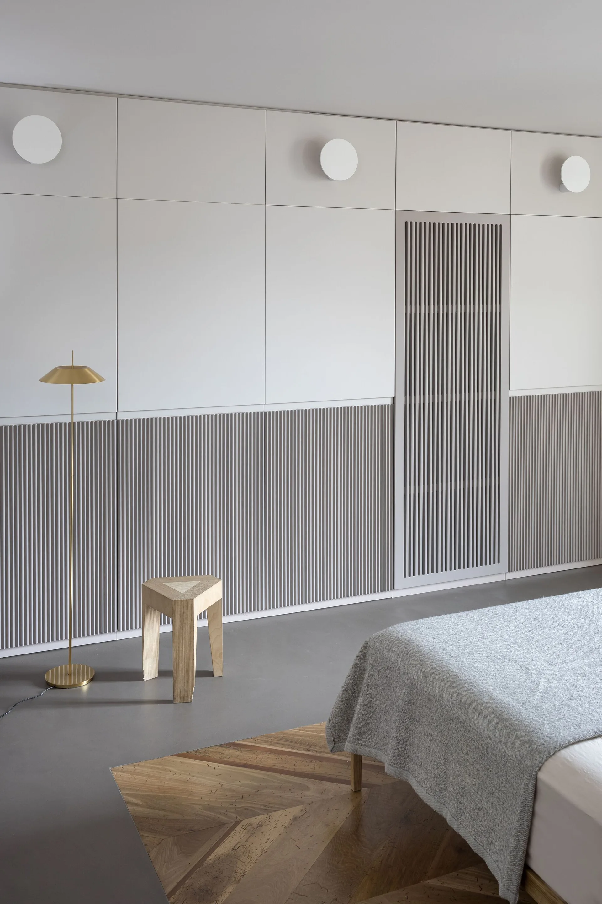 A modern bedroom features off-white and slatted grey cabinetry, a three-legged wooden stool, a thin brass lamp, and chevron wood flooring.