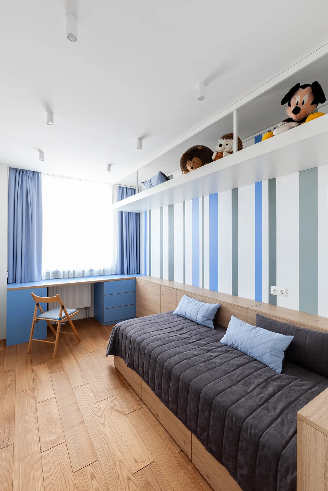 A modern kids' bedroom features a wooden daybed with built-in storage, a blue desk under the window, and blue-and-white striped wallpaper.
