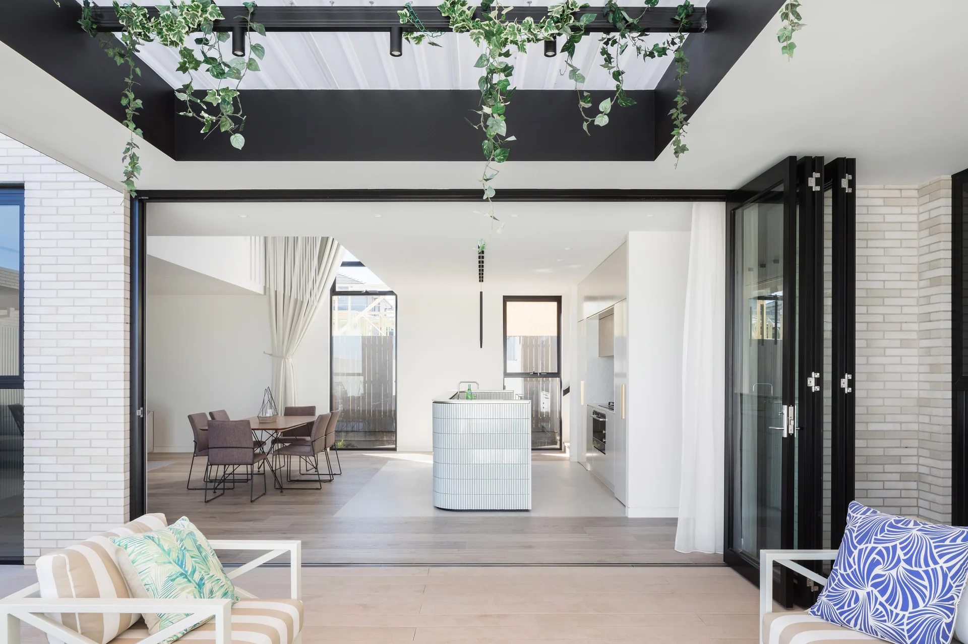 An open-plan kitchen and dining room with a curved tiled island is viewed through open black bifold patio doors.