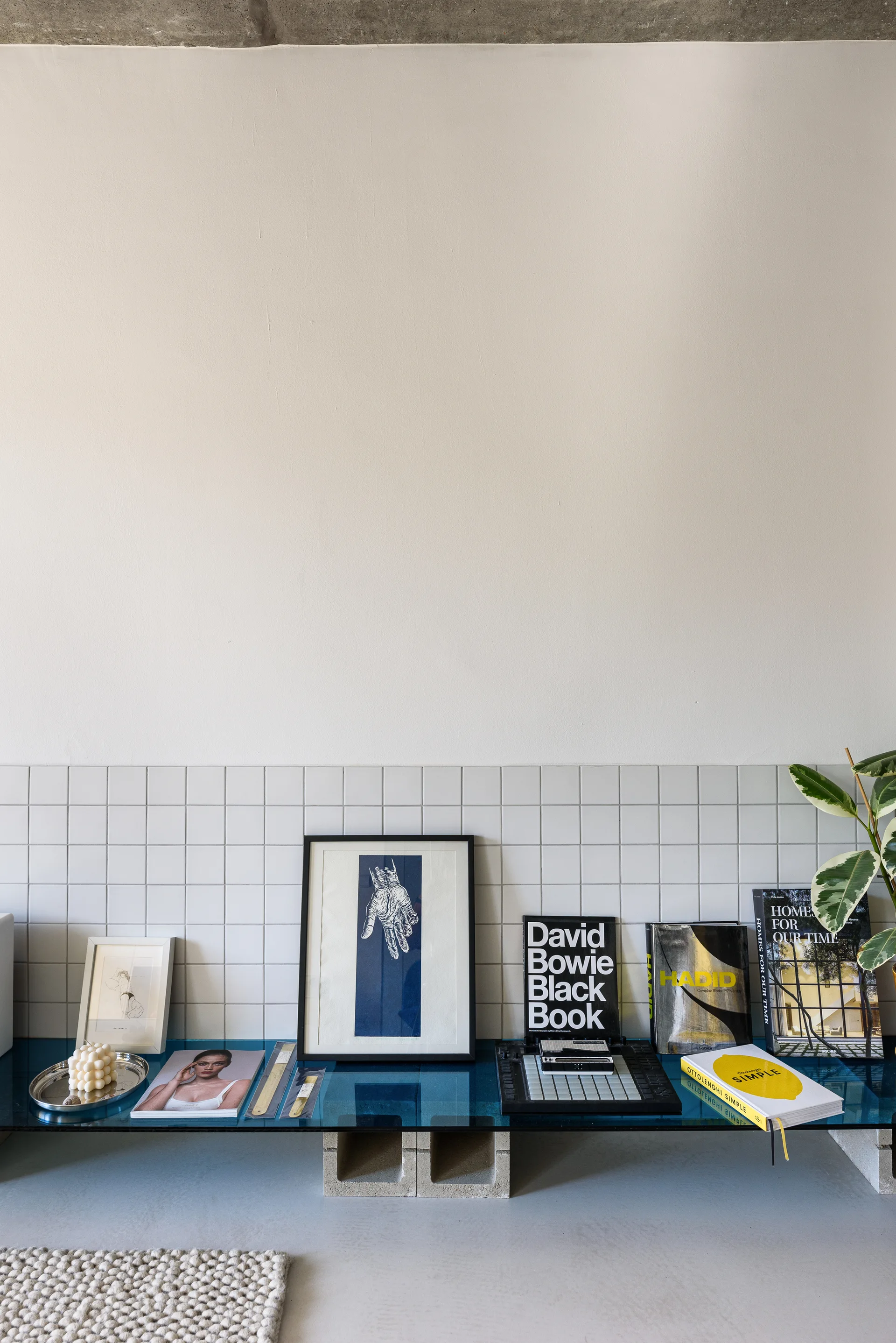 A blue glass shelf supported by cinder blocks holds books, art frames, and a candle against a white square-tiled wall.
