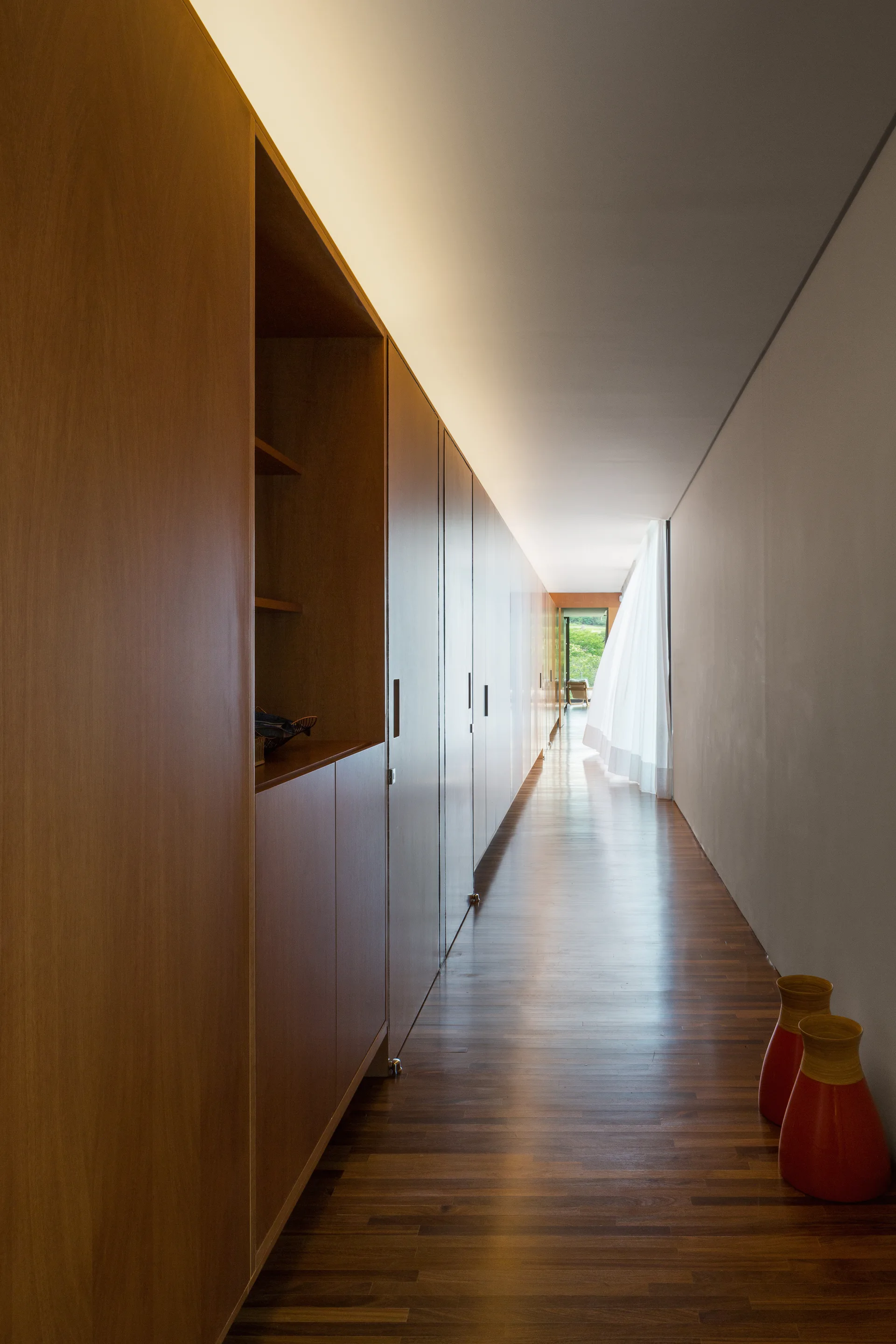 A long, minimalist hallway featuring built-in wood cabinetry, hardwood floors, a white sheer curtain, and warm cove lighting along the ceiling.