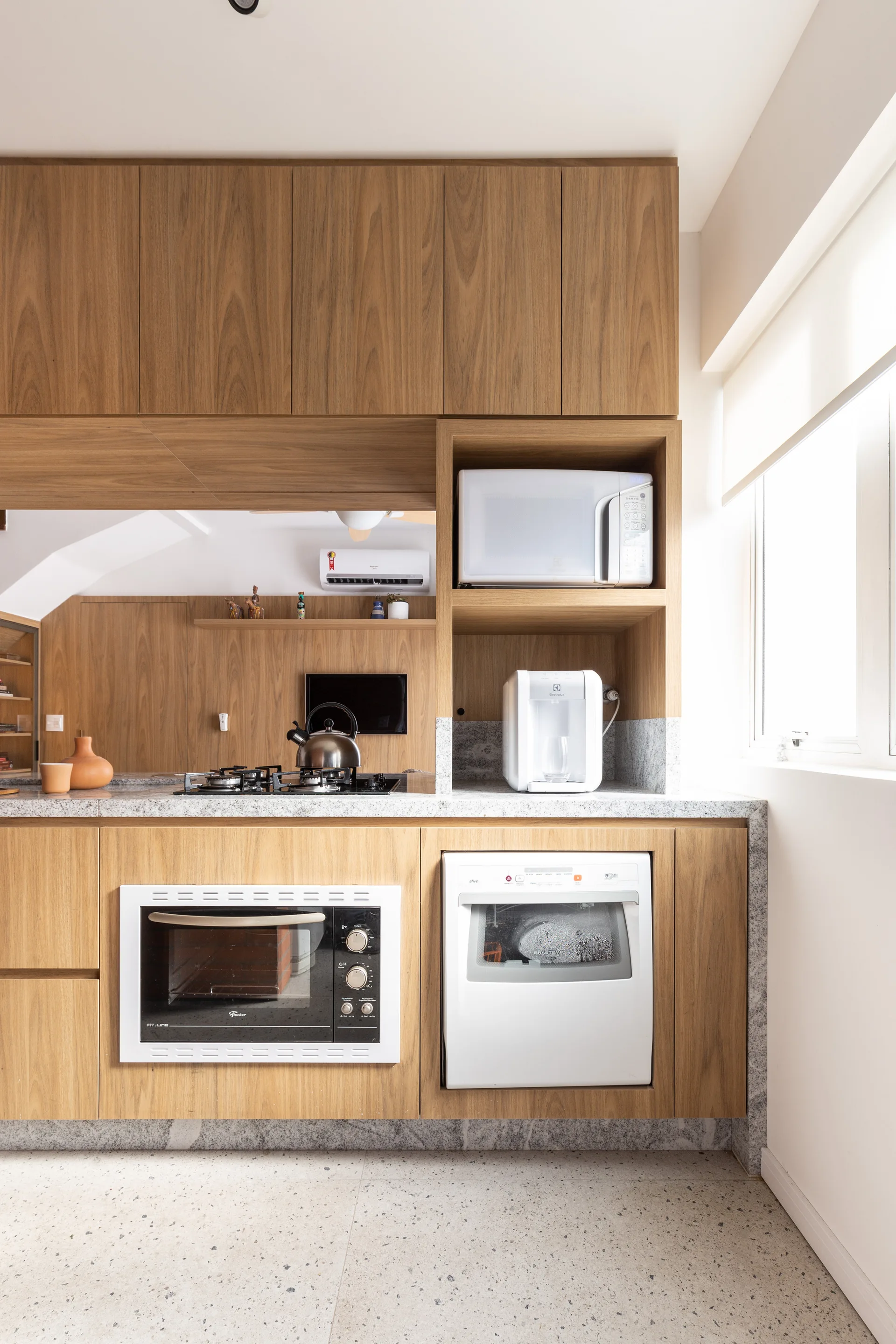 A modern kitchen featuring warm wood cabinets, grey speckled stone countertops, and built-in white appliances under natural window light.