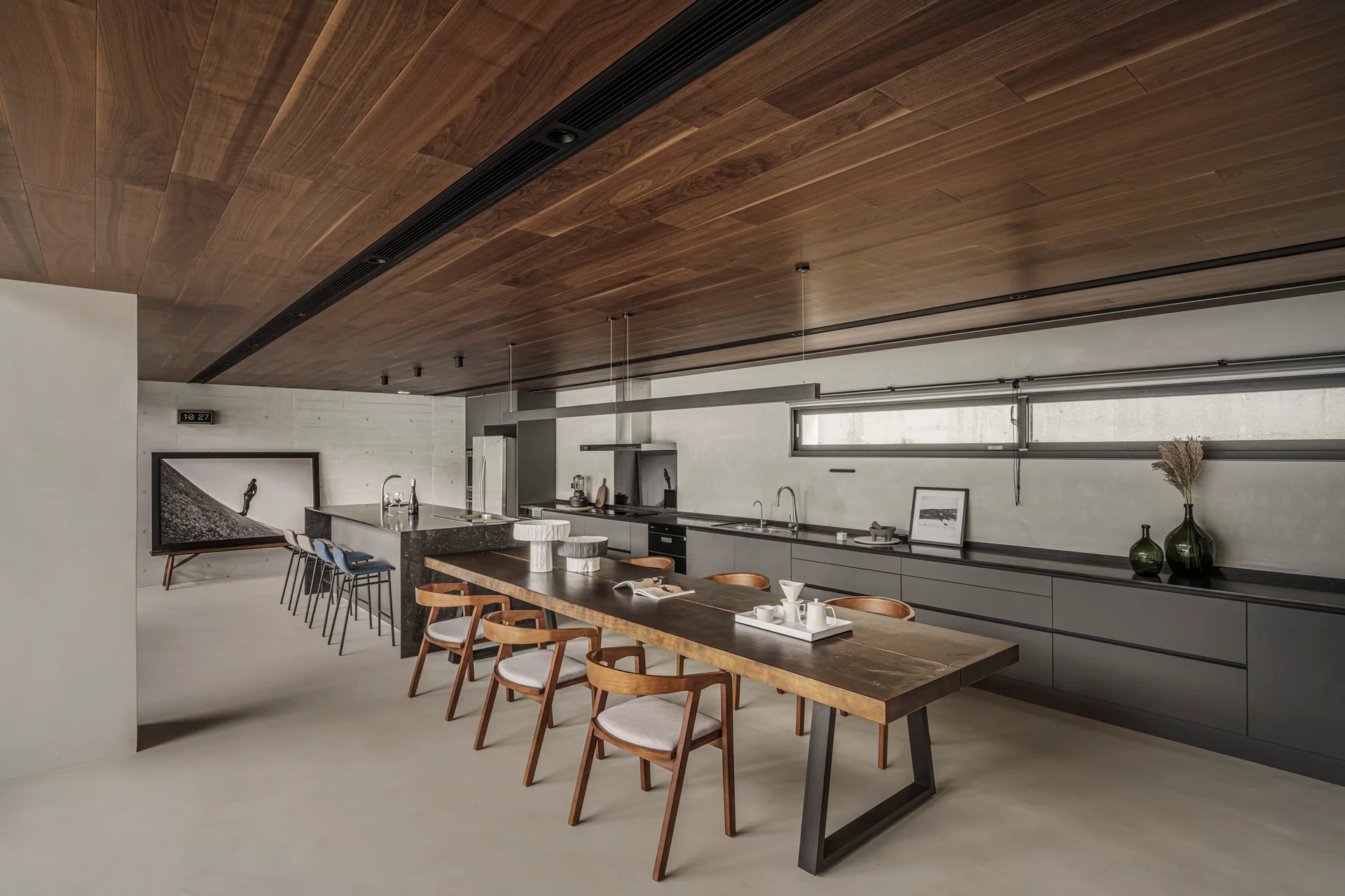 A modern kitchen and dining space with a long wooden table, dark gray cabinets, concrete walls, and a wood plank ceiling.