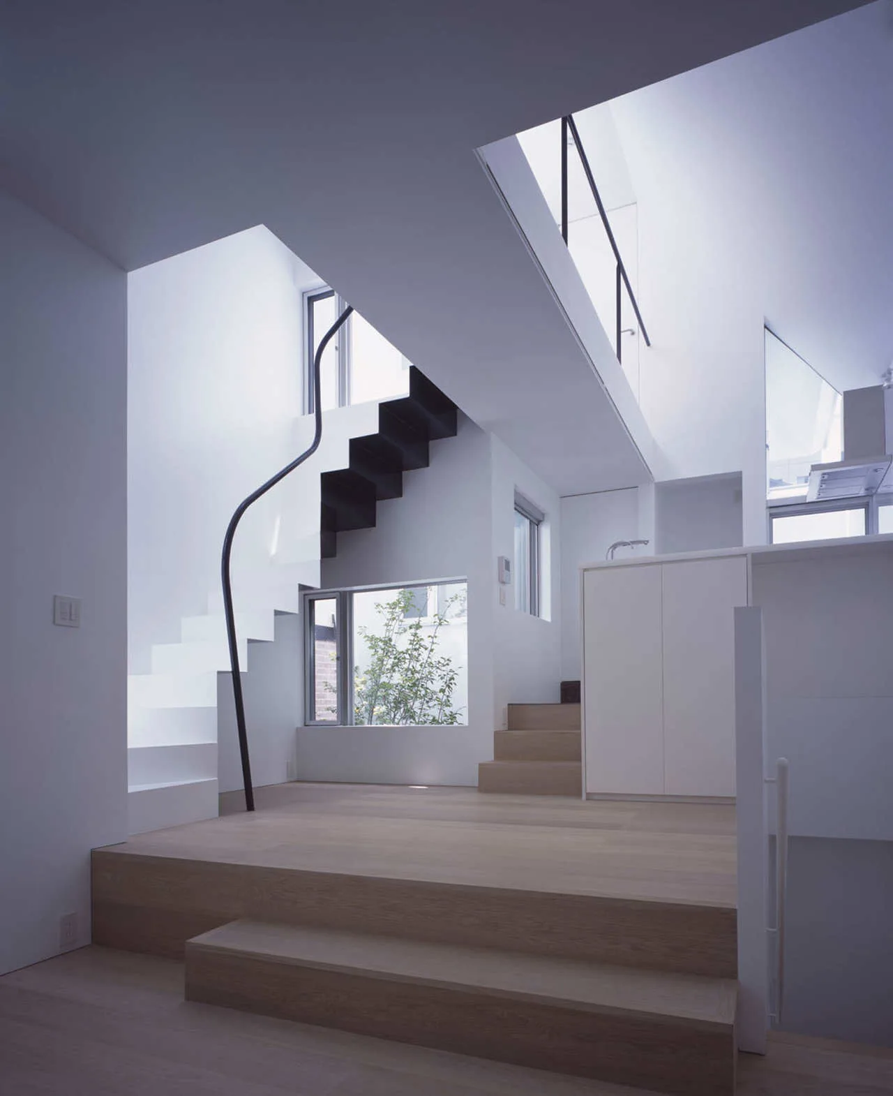 A minimalist split-level interior with white walls, light wood flooring, a floating white staircase, and a wavy black handrail beneath a skylight.