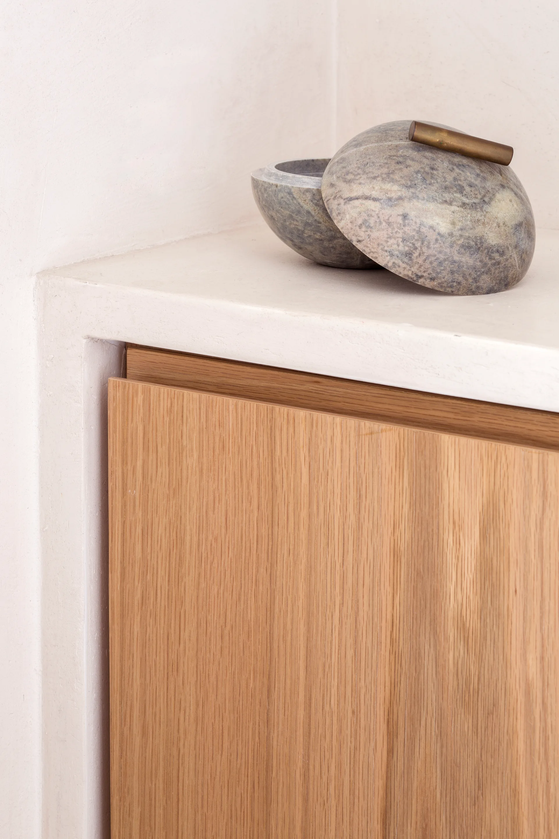 A mottled grey stone bowl with a brass handle rests on a smooth plaster shelf above a flat-front light oak cabinet.