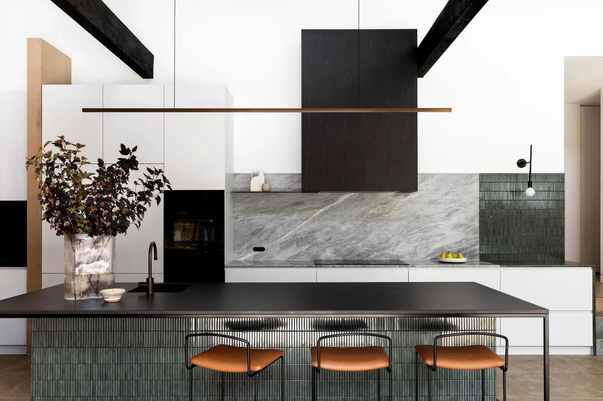 A modern kitchen with a black island clad in green fluted tile, leather-seated stools, a grey marble backsplash, and dark ceiling beams.