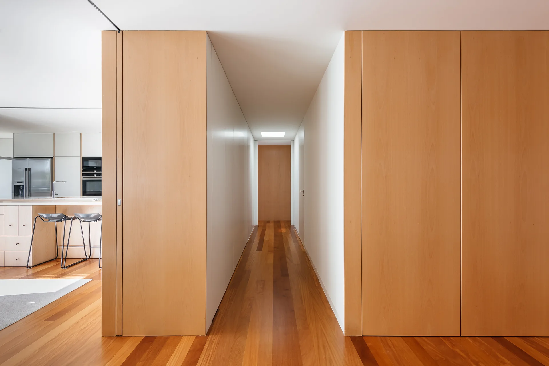 A minimalist residential hallway features warm wood plank flooring, matching floor-to-ceiling wood wall panels, and an open view into a kitchen.