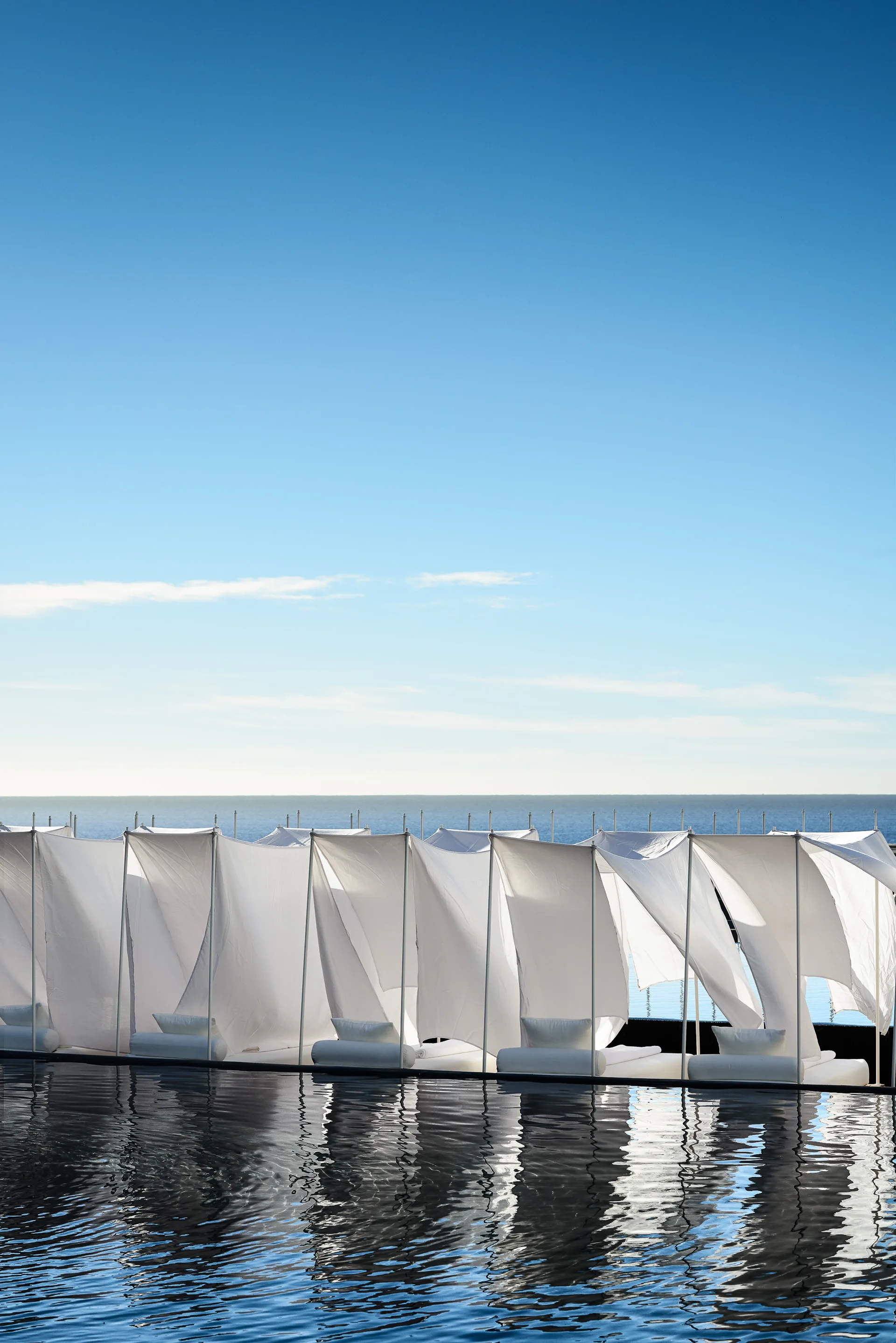 A row of white fabric poolside cabanas sits next to a reflective dark swimming pool under a clear blue sky.