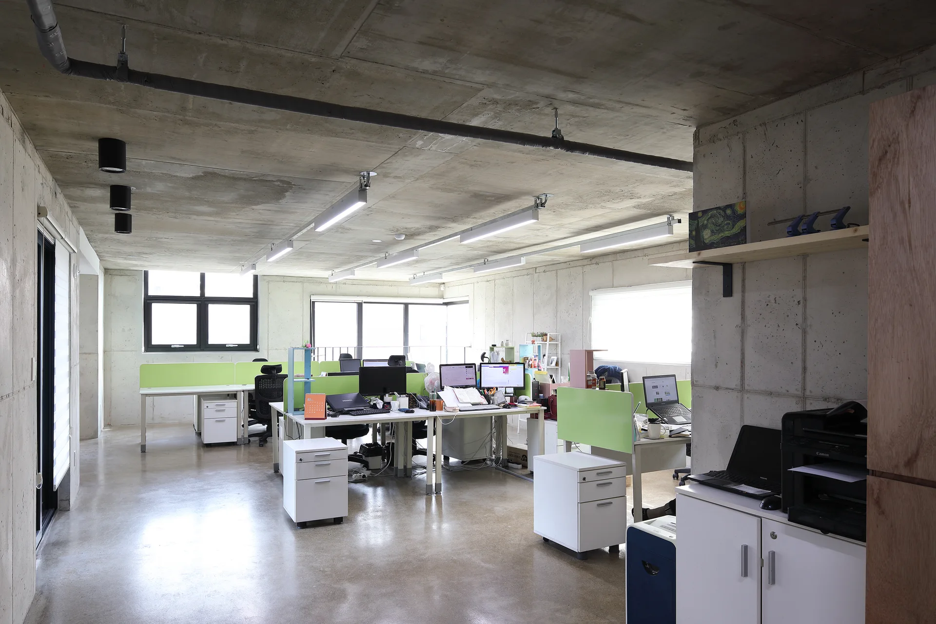 An open-plan office features exposed concrete walls and ceilings, white desks with lime green privacy dividers, and black mesh chairs.