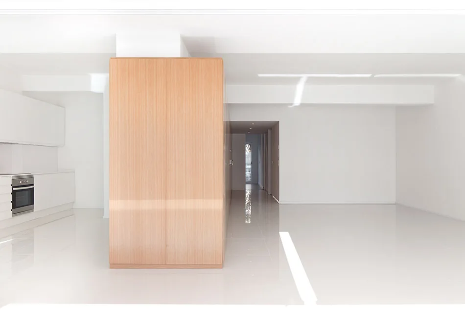 A minimalist white kitchen and living space features a large light wood cabinet partition on a highly reflective glossy tile floor.