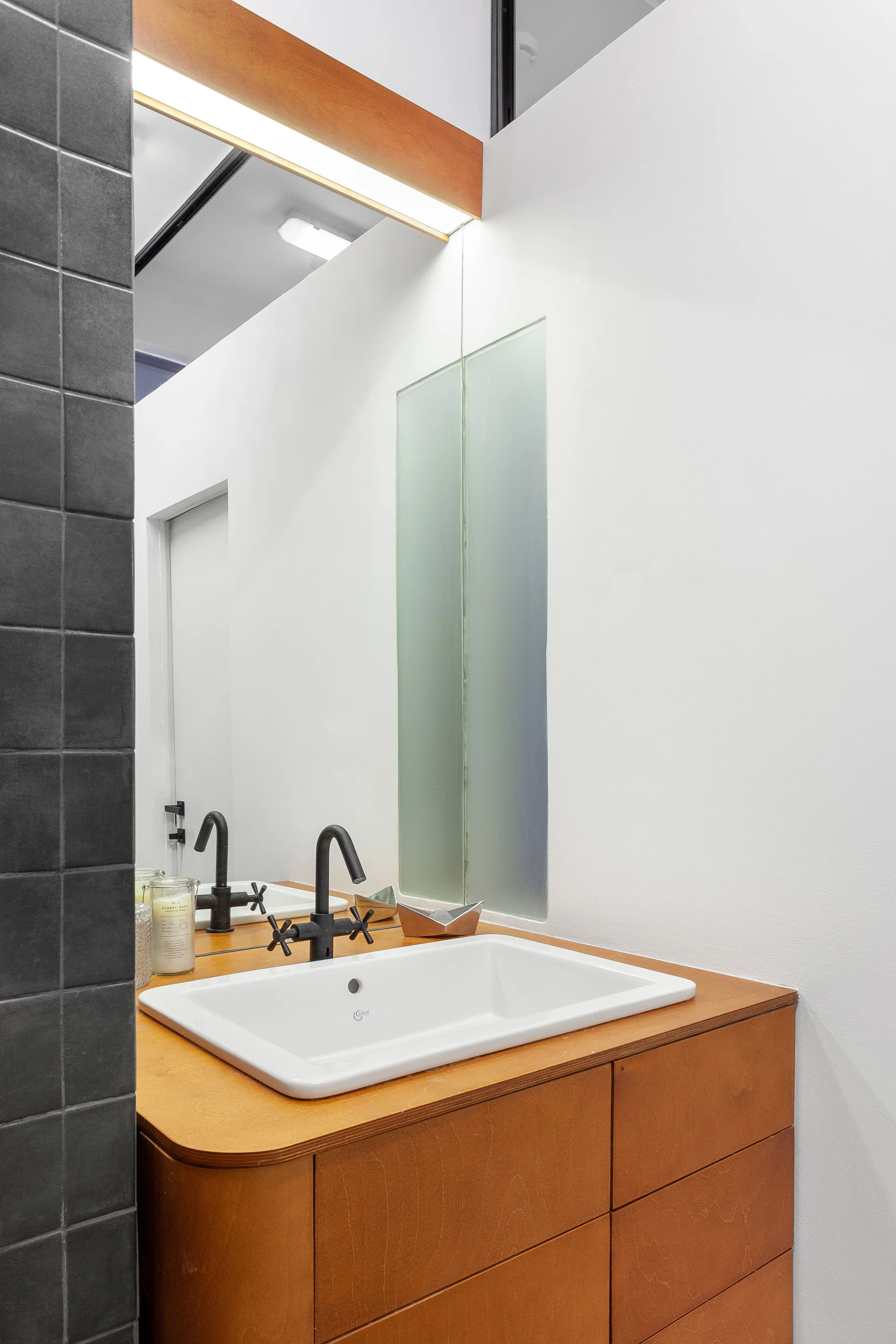 A wood-toned bathroom vanity with a white ceramic sink and matte black faucet stands next to a dark grey tiled wall beneath a large mirror.