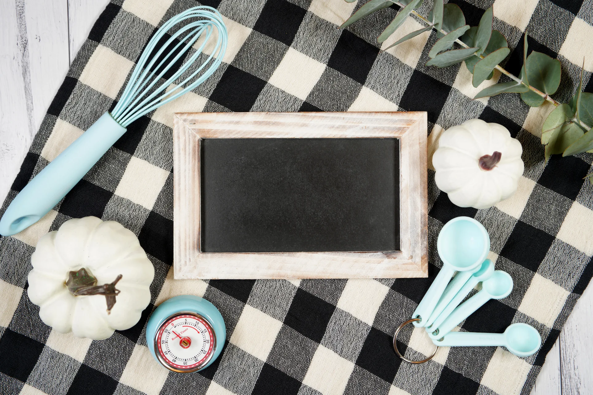 A small chalkboard, light blue kitchen tools, and white decorative pumpkins are arranged on a black and white buffalo check cloth.