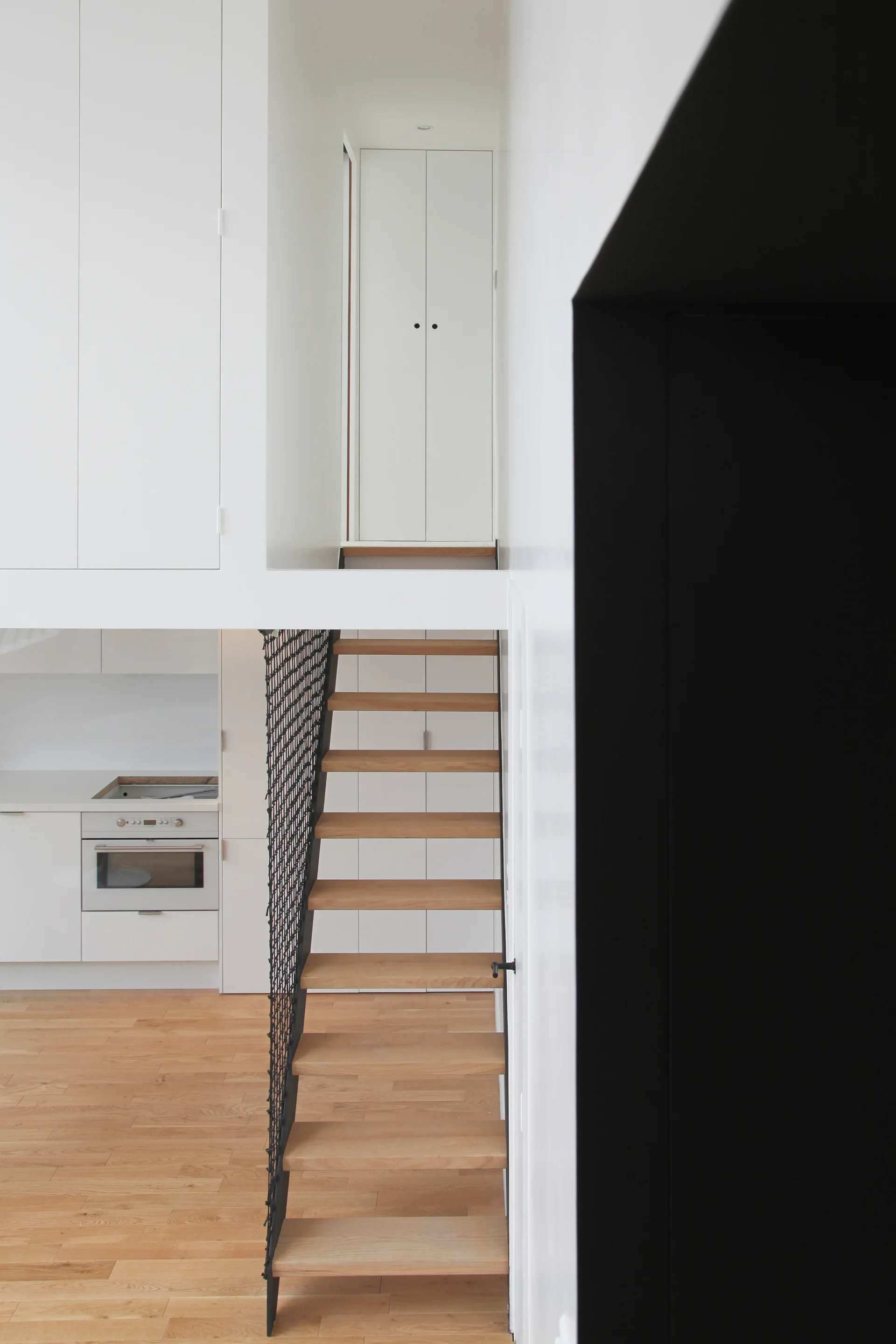 An open-riser wooden staircase with a black mesh railing stands in a minimalist interior featuring white built-in cabinetry and a kitchen background.
