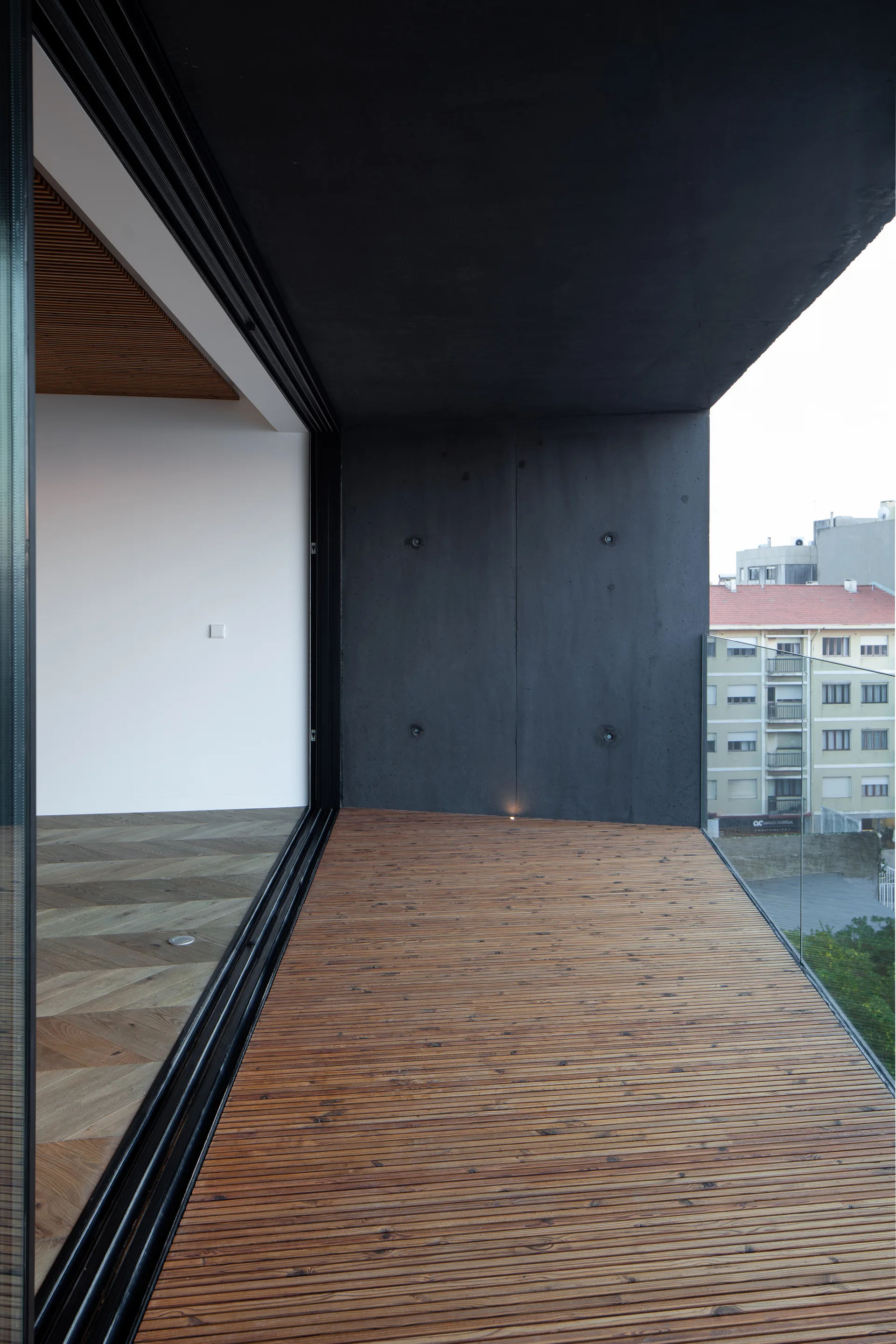 A modern balcony features warm wood slatted decking, a dark exposed concrete wall, a frameless glass railing, and sliding doors opening to an indoor room.