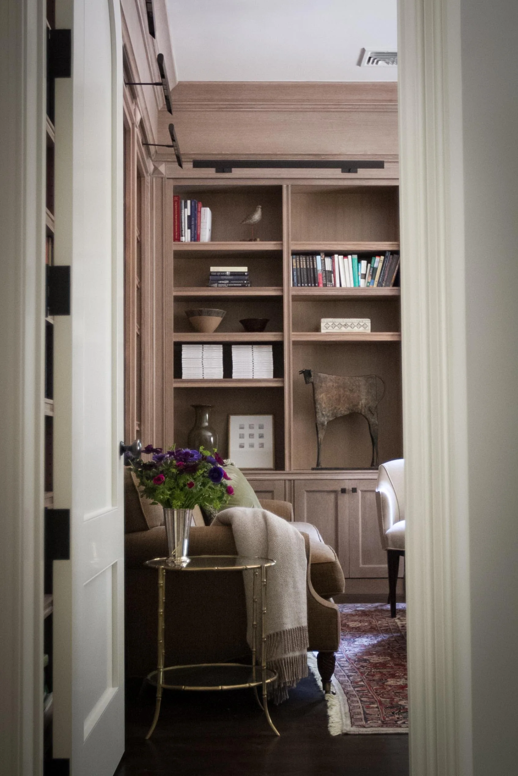 A view through a white doorway into a traditional home library featuring floor-to-ceiling wood bookshelves, an armchair, and a patterned rug.