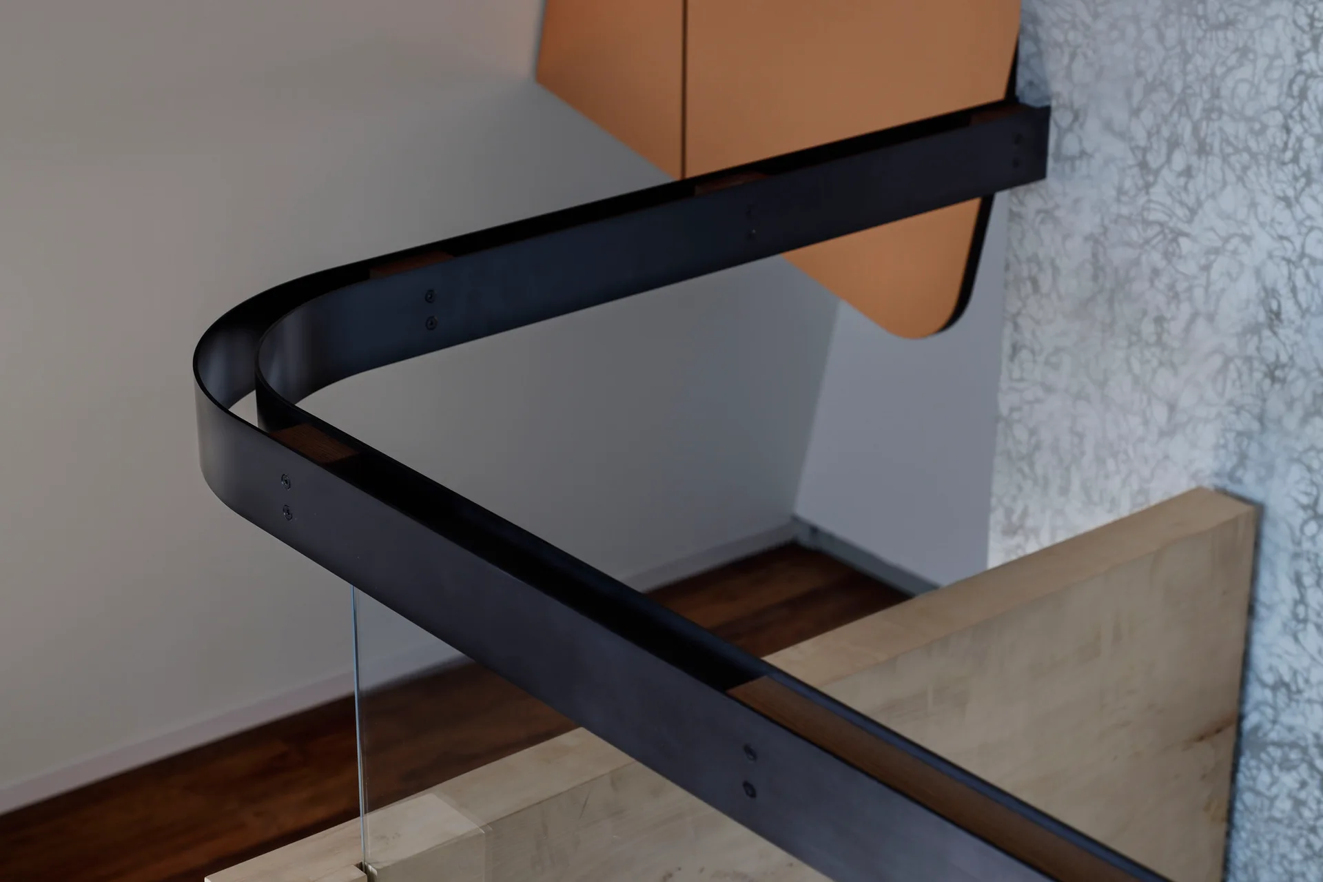Close-up of a modern matte black metal handrail curving over a clear glass panel and light wood stair base.